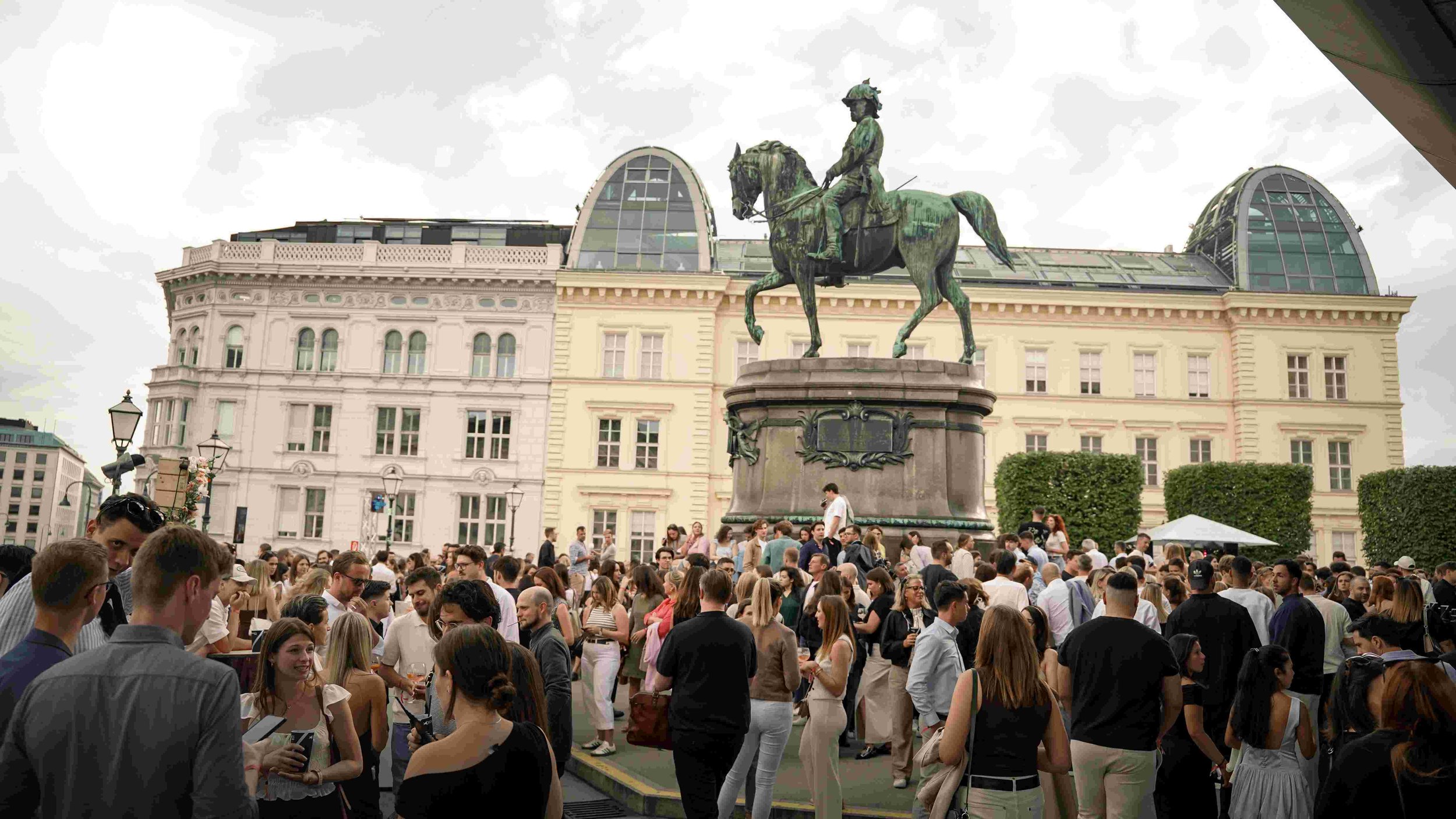 A large crowd of people gathered around a statue of a man on horseback in a city square. The background features historic buildings and a cloudy sky.