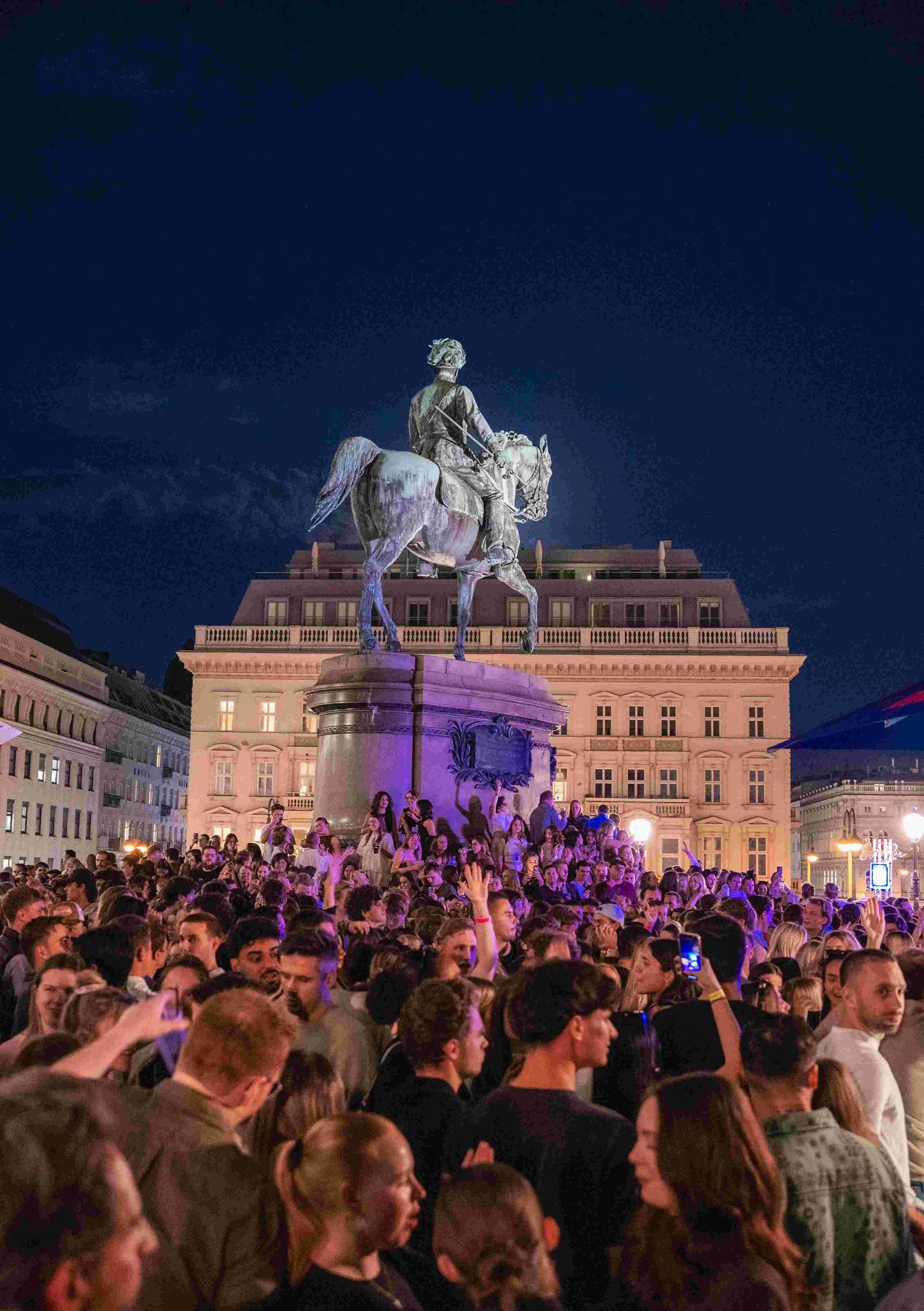 Eiffel Tower replica statue at night with a large crowd gathered around, city buildings in the background, illuminated with pink and purple lights.