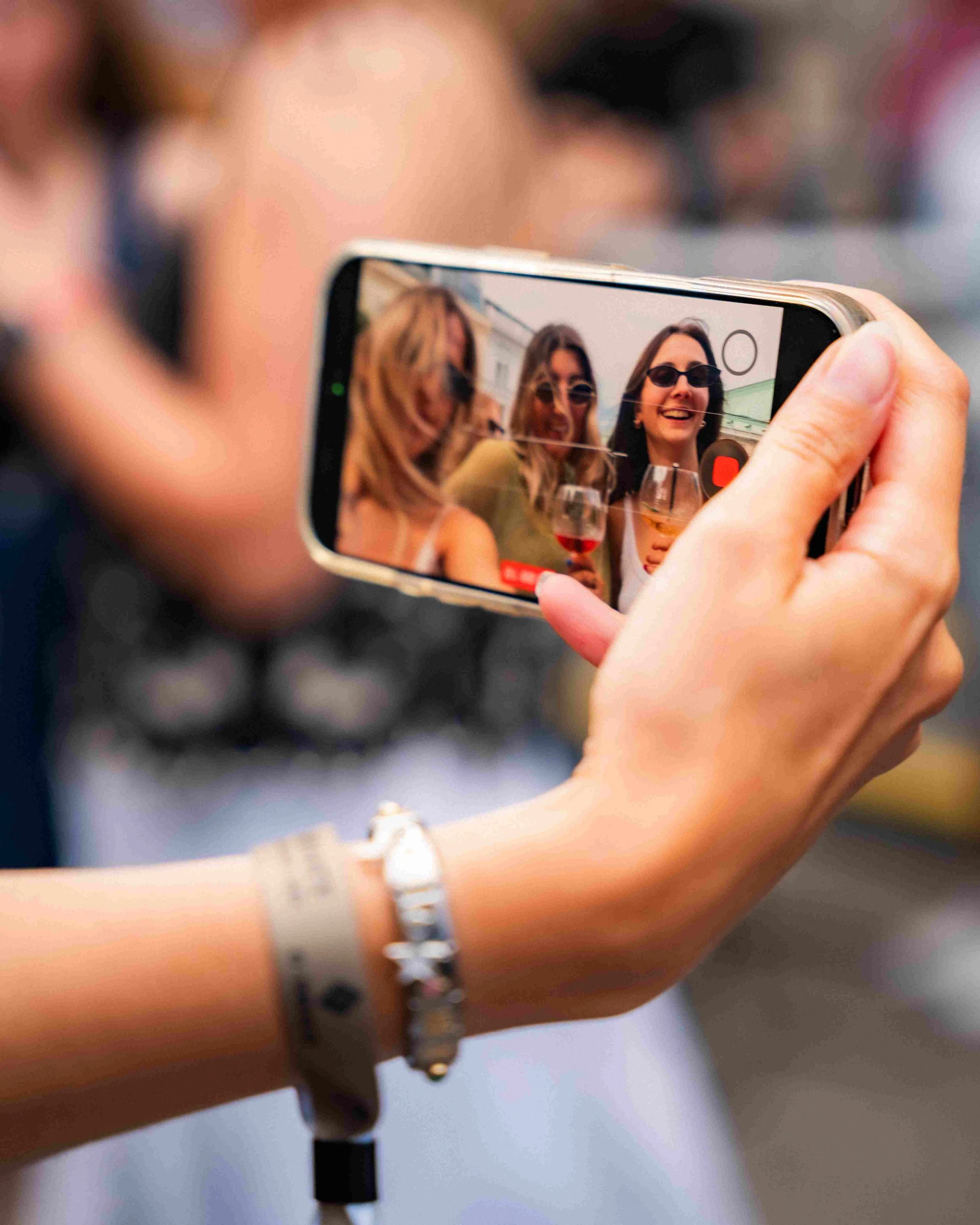 A person holding a smartphone taking a photo of three women at an outdoor gathering, with wine glasses, all wearing sunglasses and smiling.