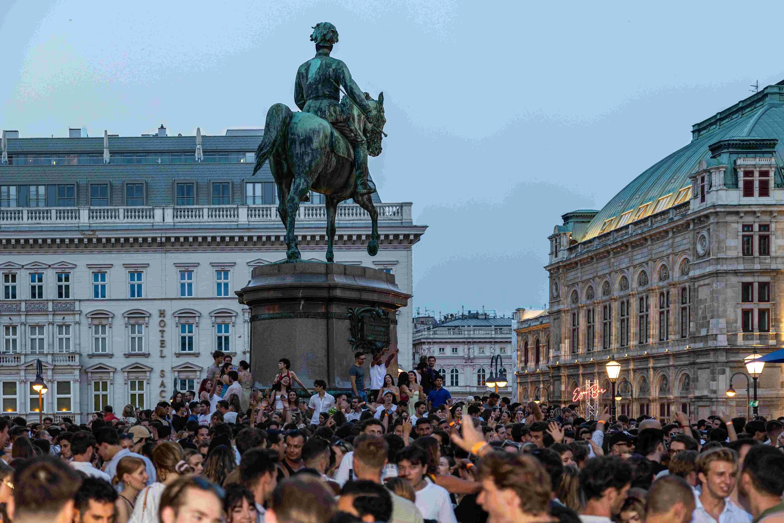 A large crowd of people gathered in a public square with a bronze equestrian statue on a stone pedestal in the background.