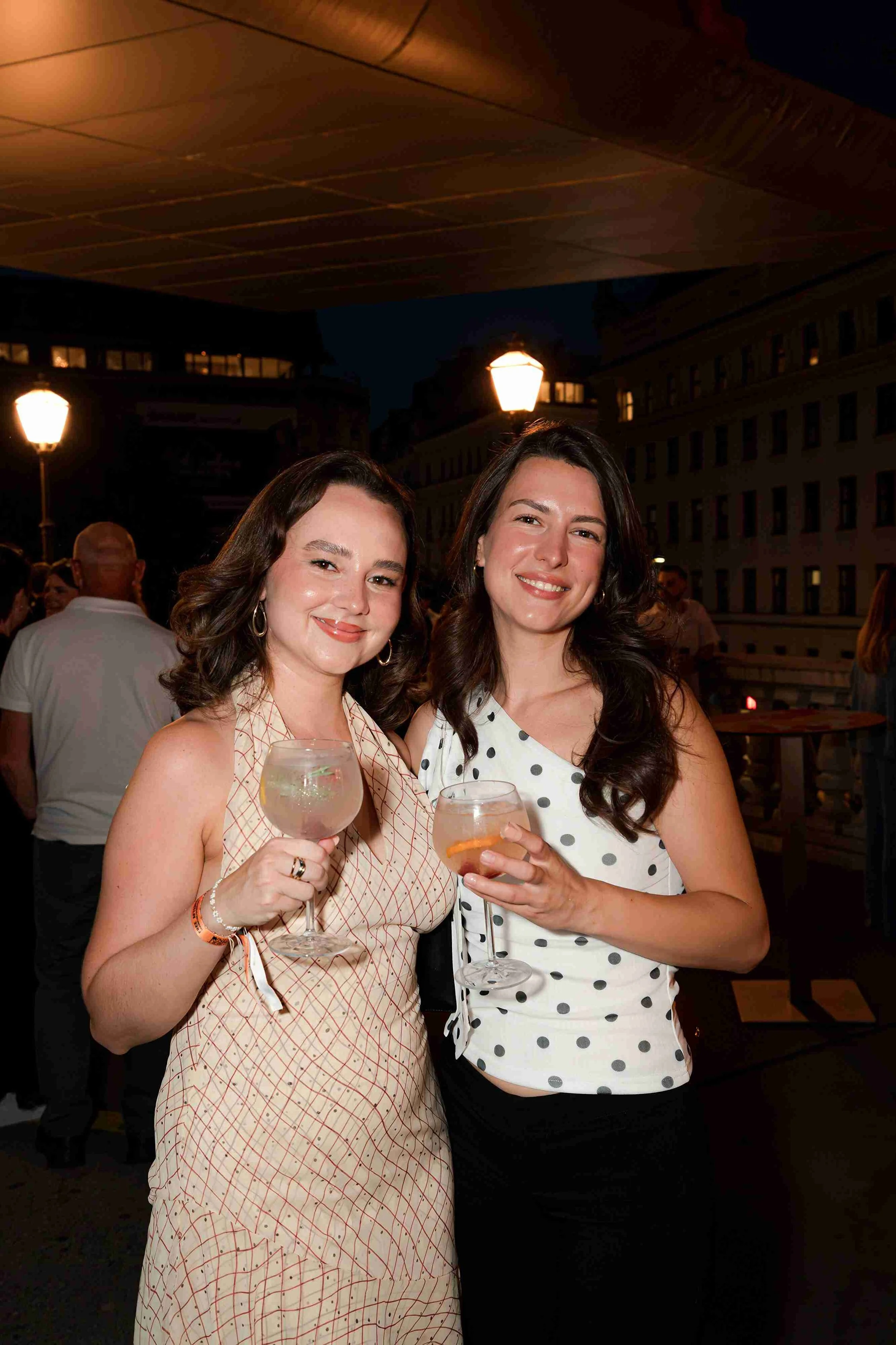 Two women smiling and holding drinks at an outdoor evening gathering, with city buildings and streetlights in the background.