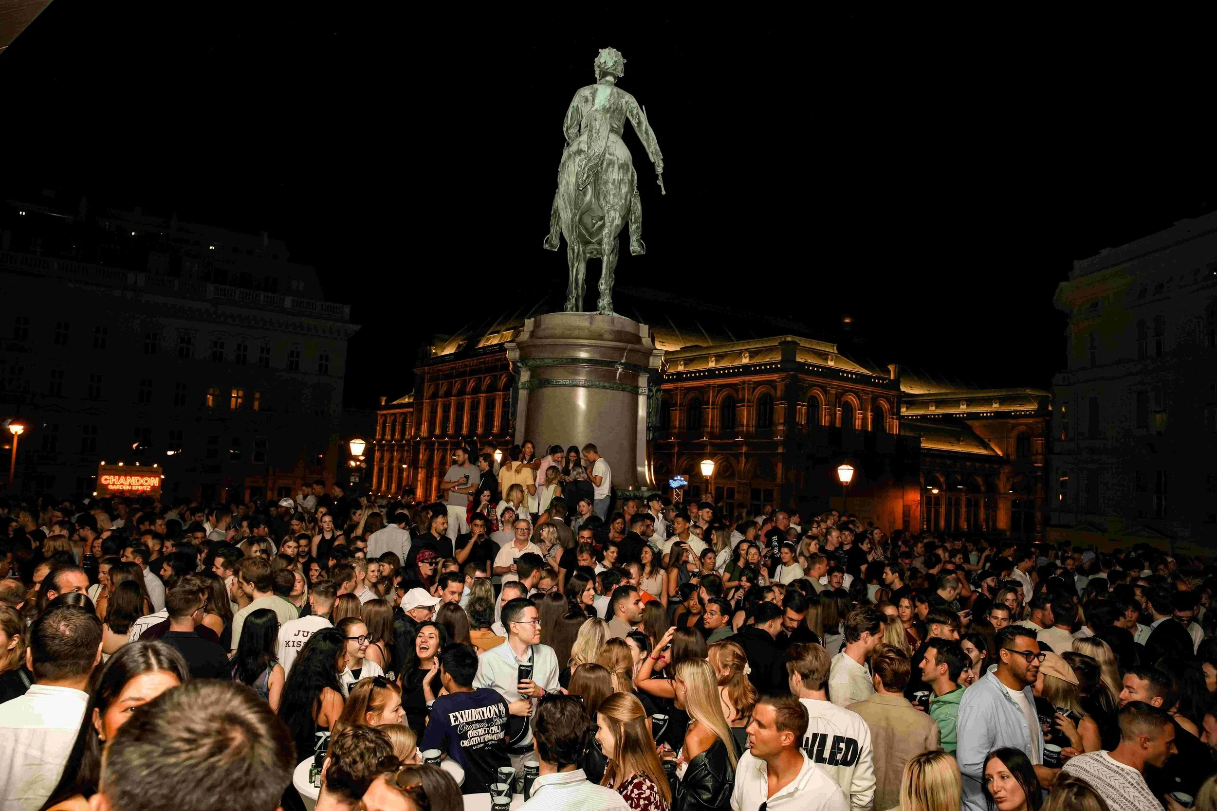 Crowd of people gathered in front of a statue of a man on horseback at night, with lit buildings in the background.
