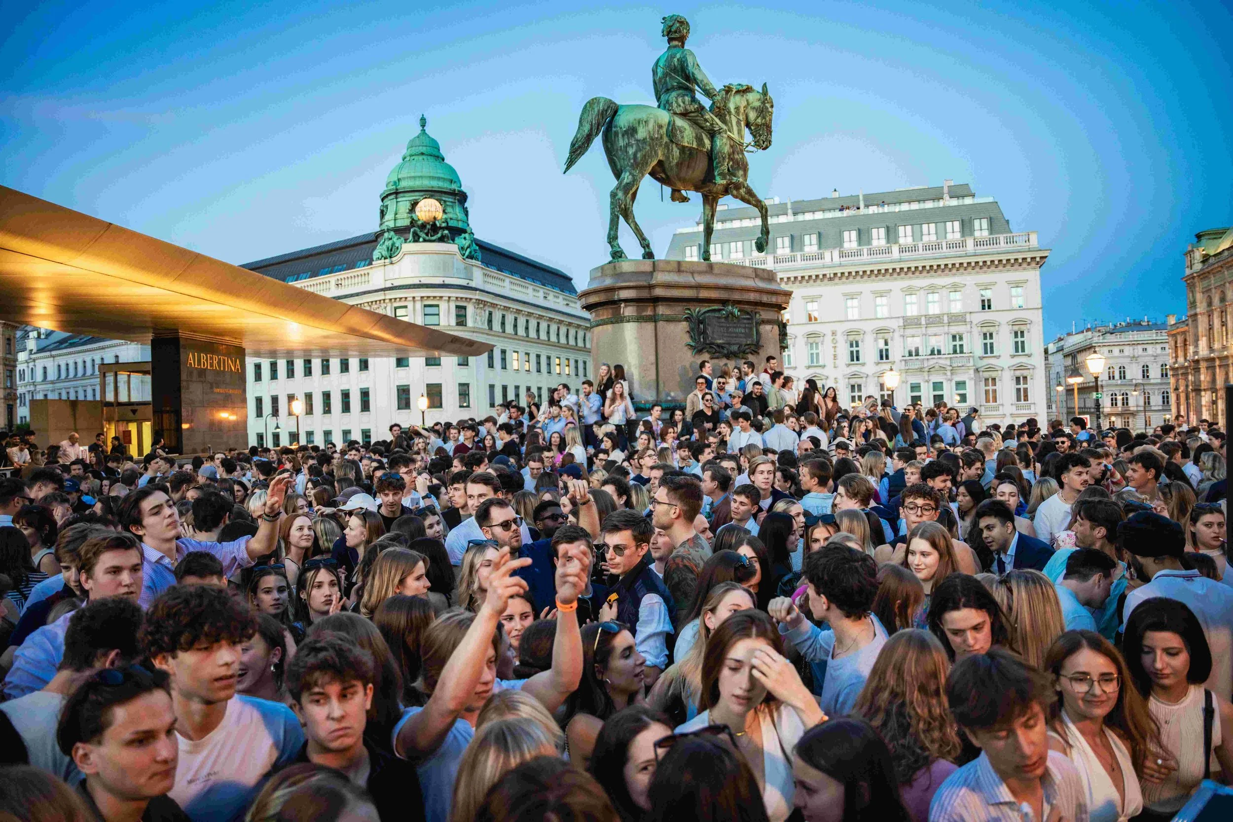 Large crowd of people gathered in a city square with an equestrian statue and historic buildings in the background.