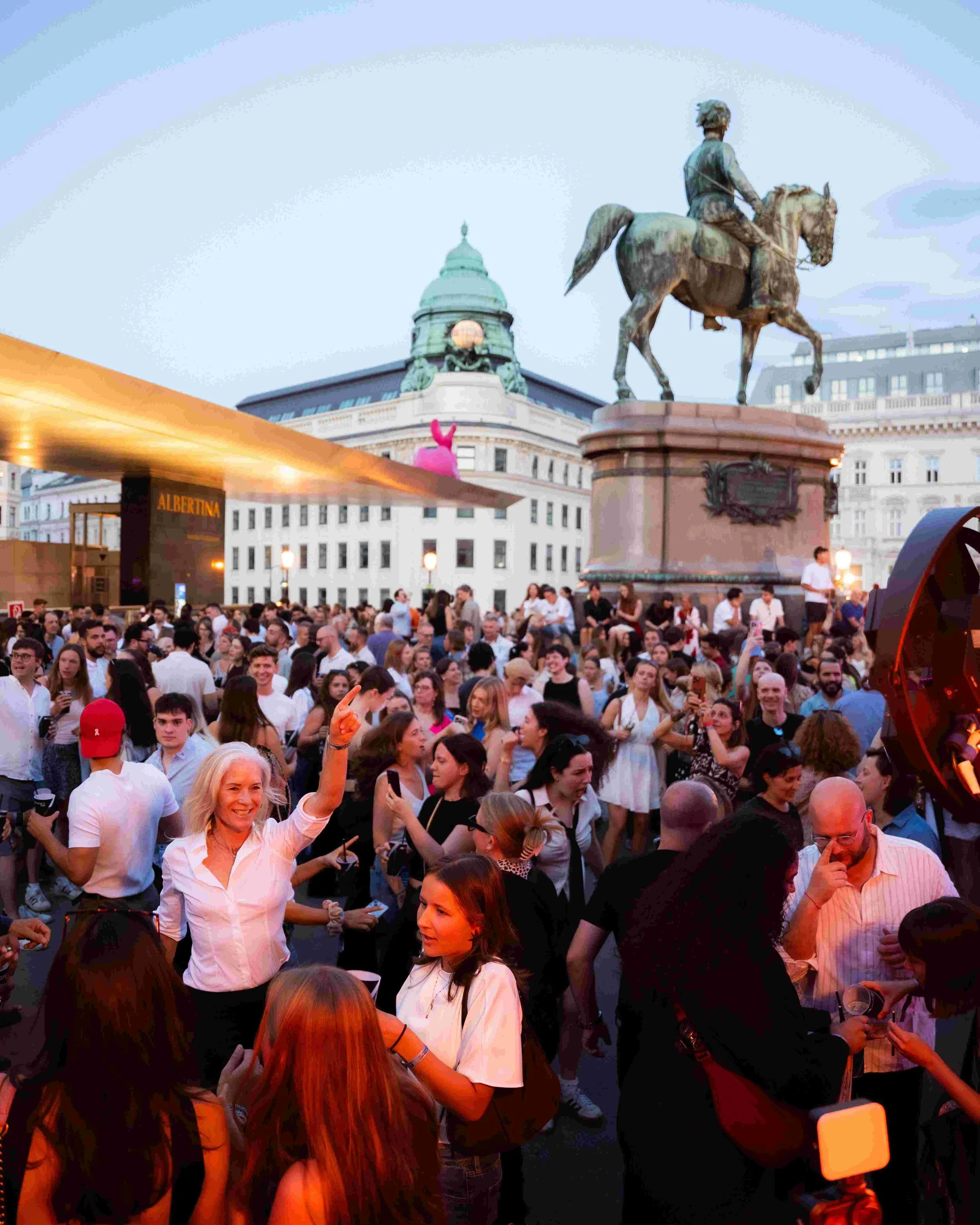 A large crowd of people gathered outdoors with a historic building and monument in the background. Some people are smiling and taking photos.