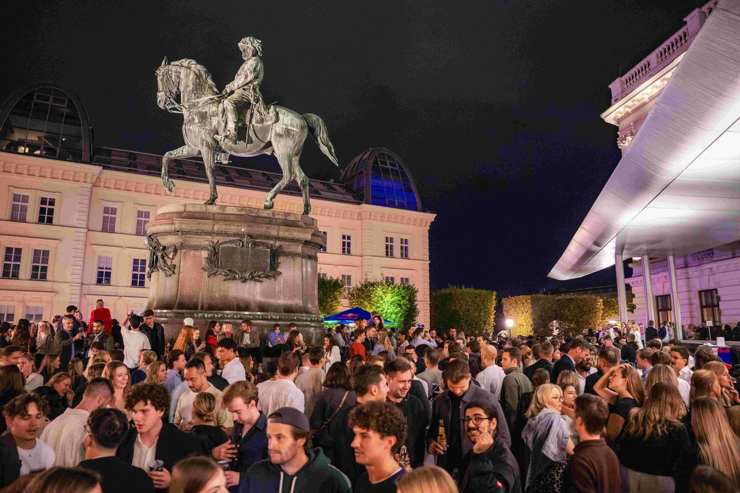 A large crowd of people gathered at night in front of a historic building with a statue of a man on a horse. The building is illuminated with colorful lights, and there is a modern glass structure with a curved roof on the right side of the image.