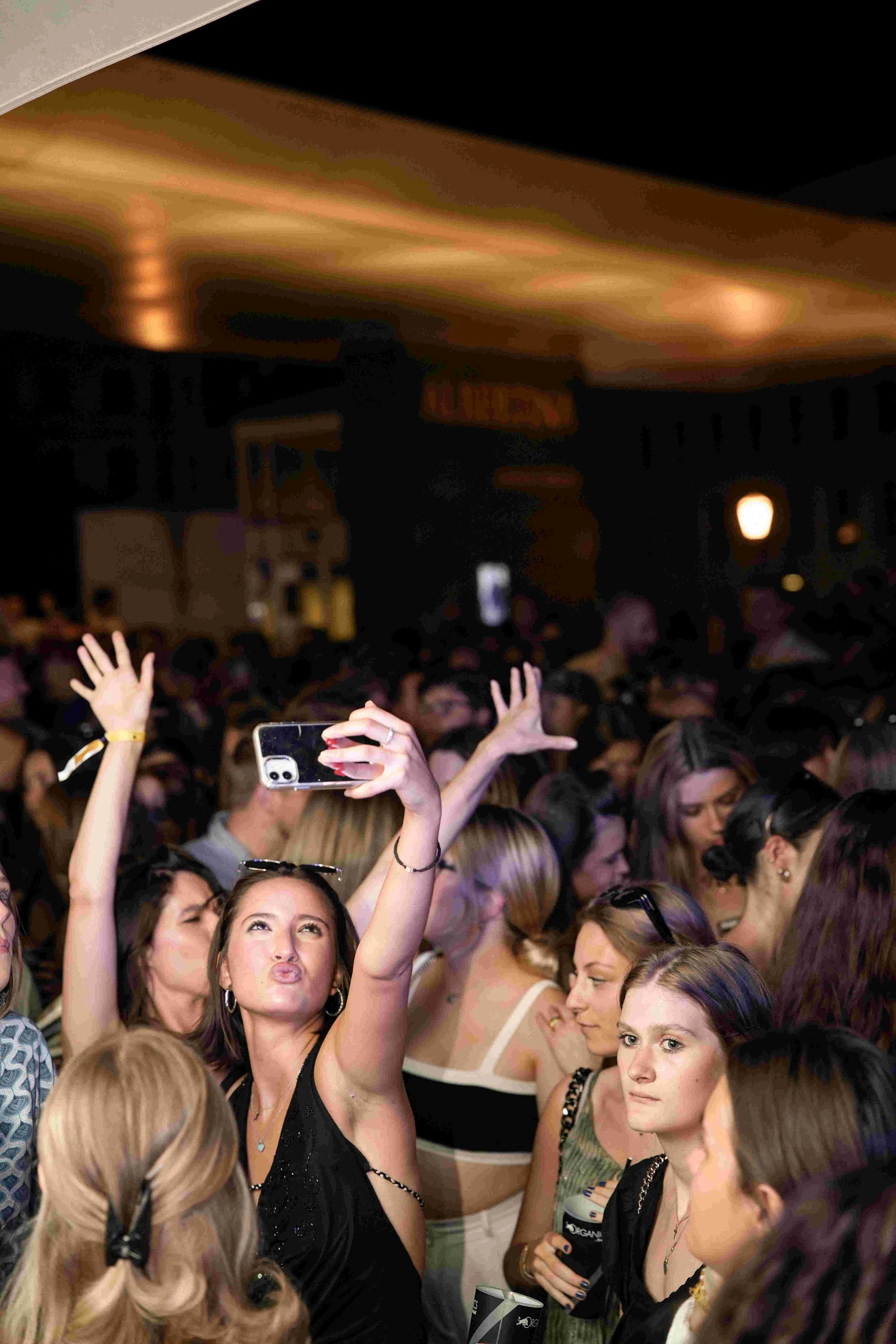 Crowd of women at a lively indoor event, many raising their hands and taking photos, with some holding drinks and wearing casual to semi-formal attire.