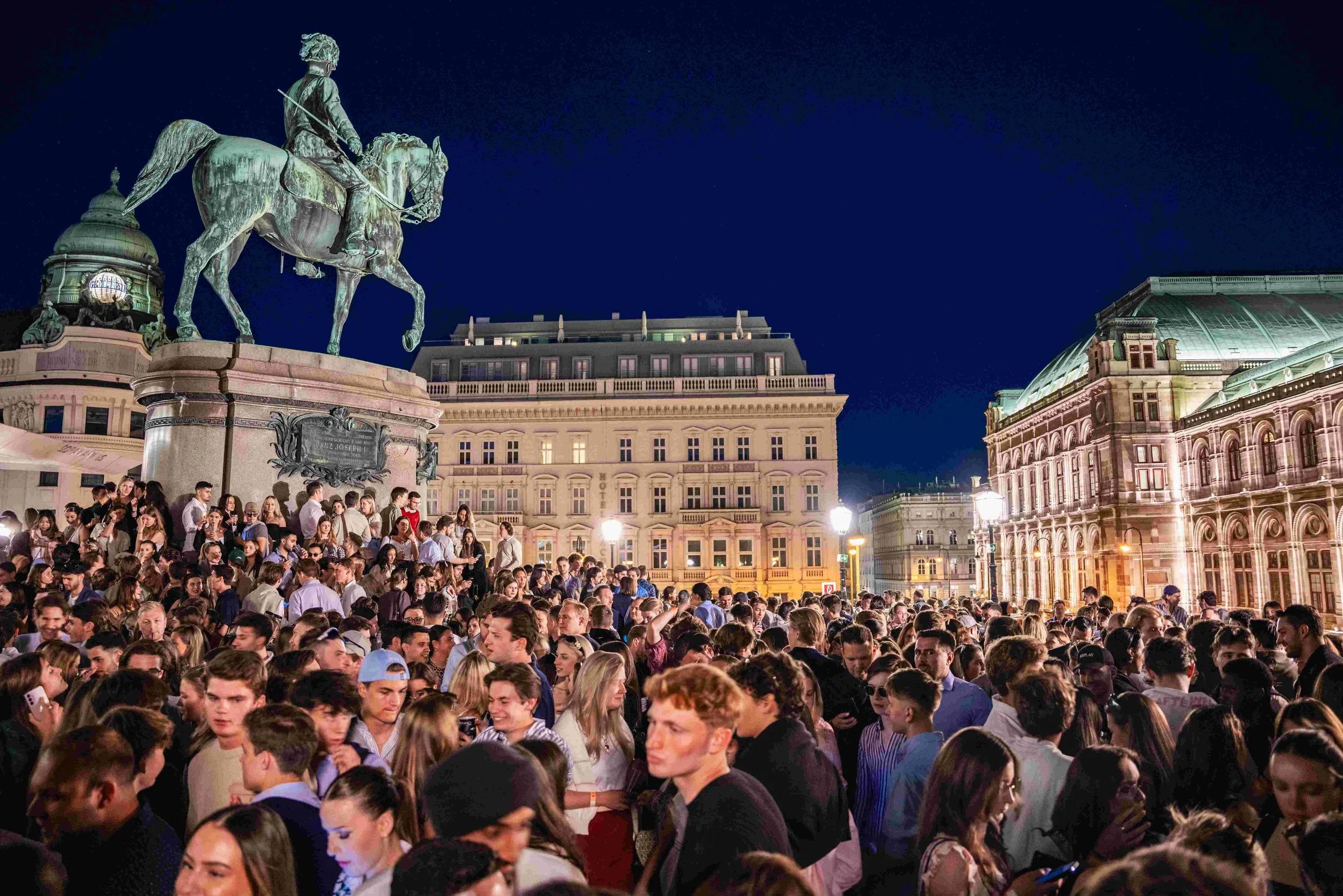 A large crowd of people gathered in an outdoor city square at night, with historic buildings and a large equestrian statue of a man in armor on a horse visible in the background.