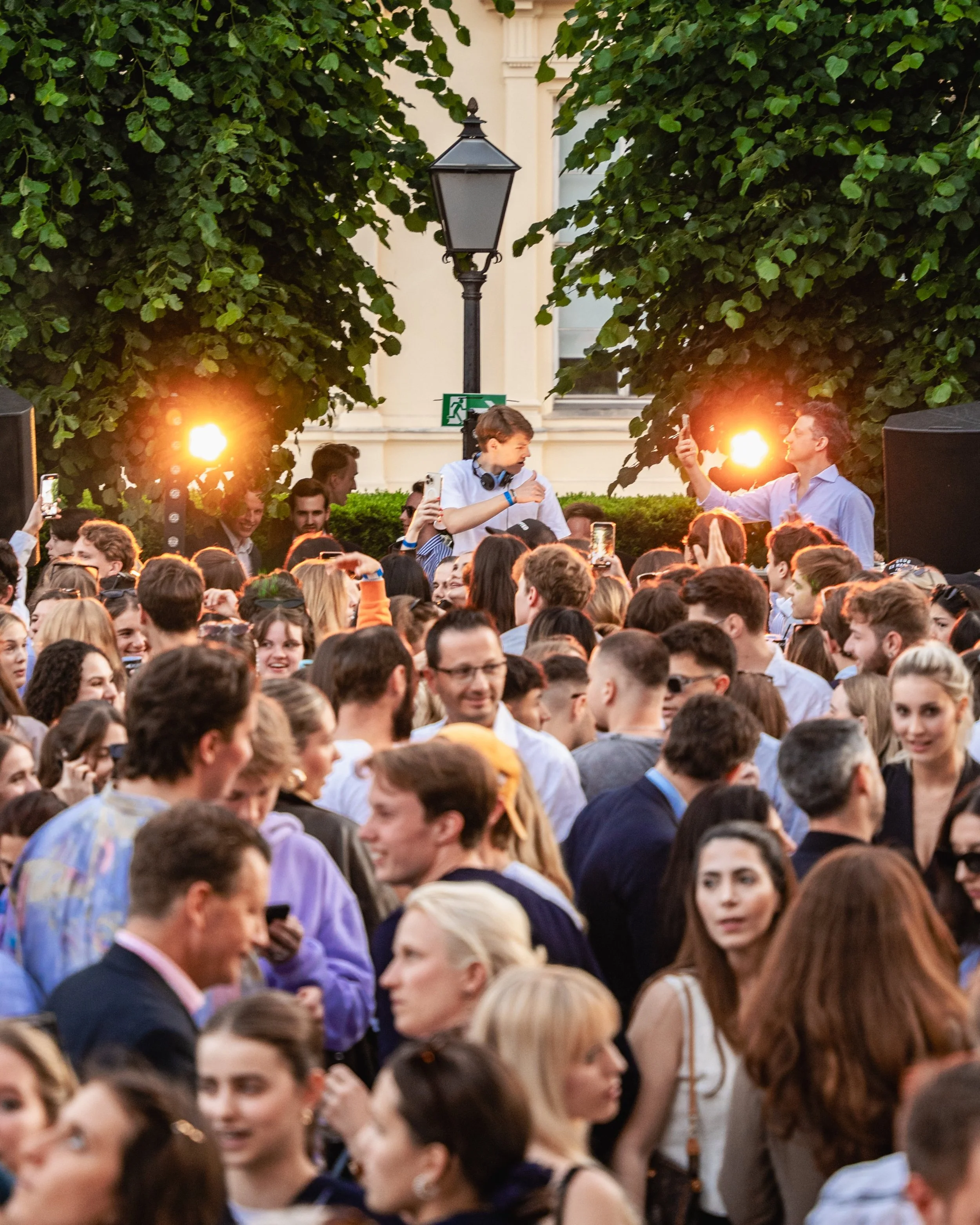 Crowd of people gathered outdoors during evening, with a background of trees and a building, illuminated by warm lights.