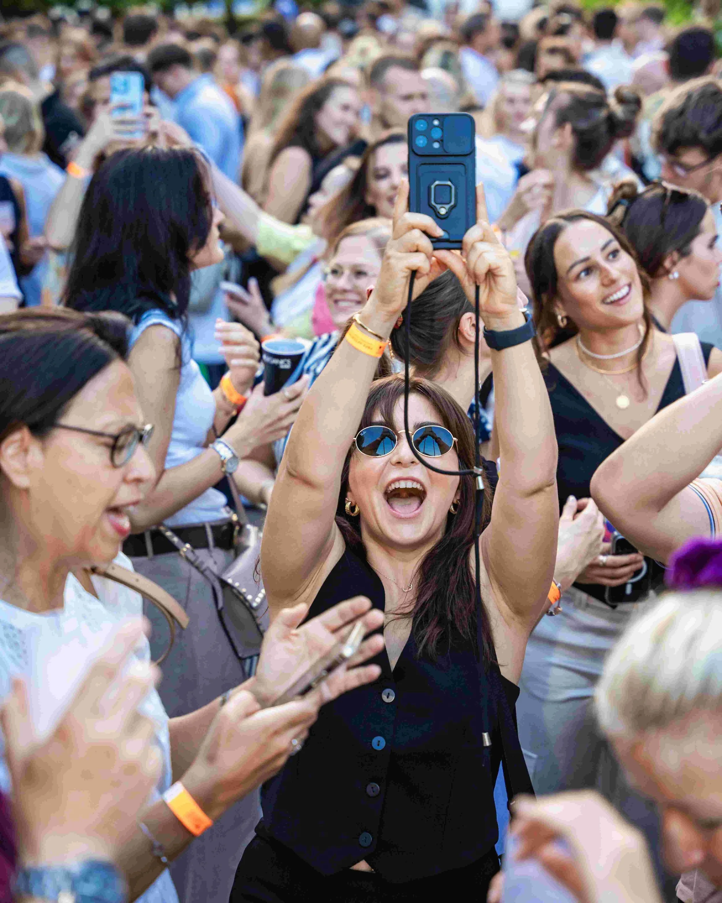 A crowd of people gathered outdoors, with one woman in center taking a selfie with her phone. The people are smiling, talking, and seem to be enjoying a social event.