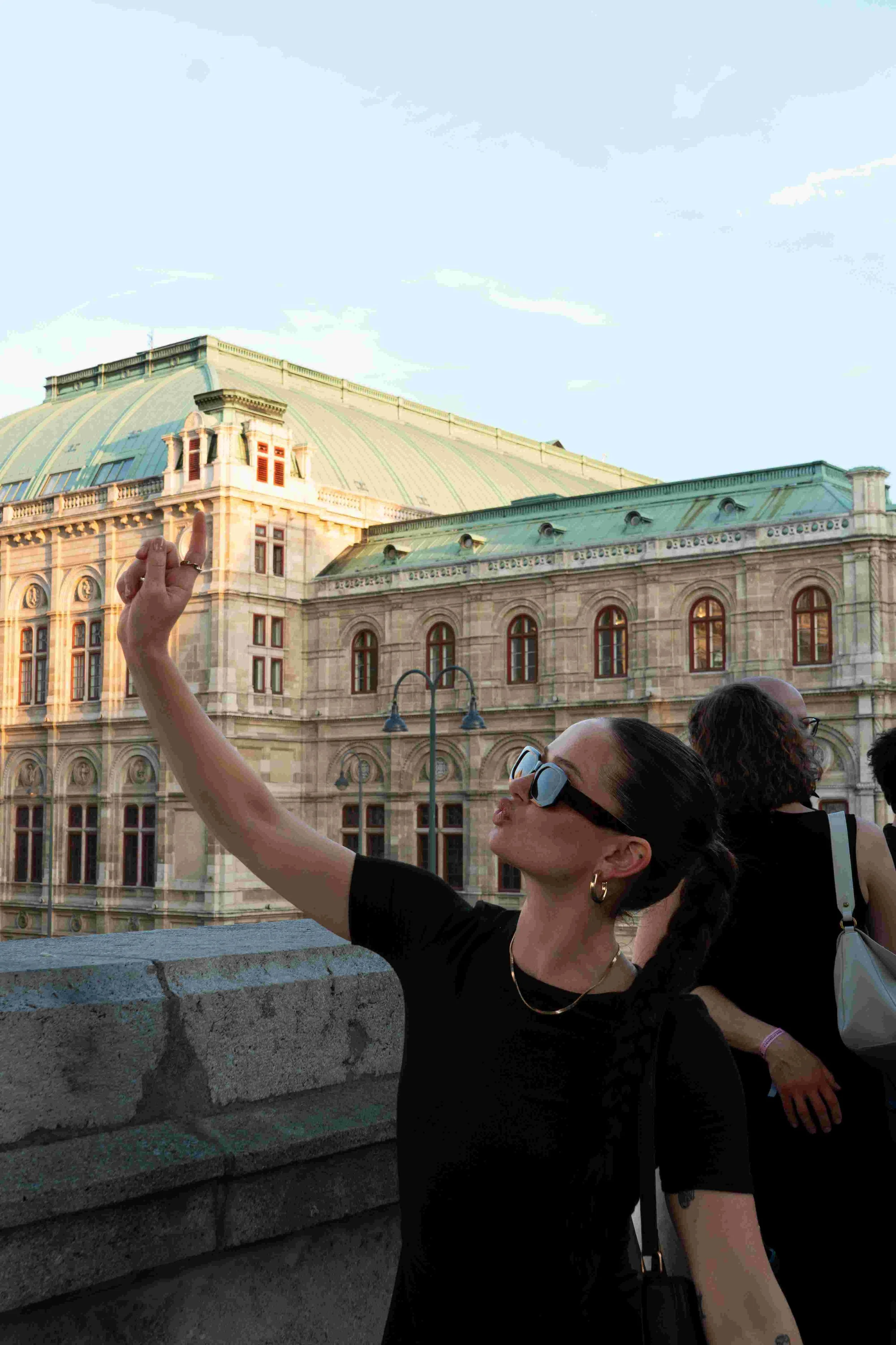 A woman in black taking a selfie with a historic building in the background, under a blue sky with some clouds.