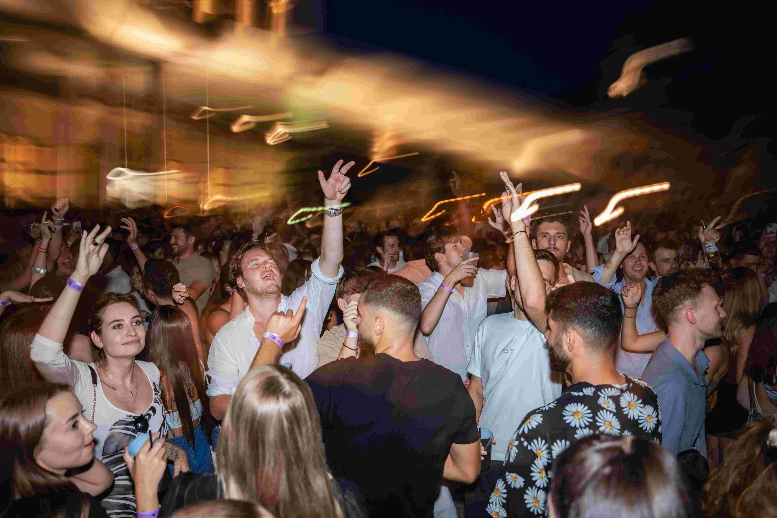 Crowd of people at a lively event or concert at night, with some raising their hands and enjoying the moment under dynamic lighting.