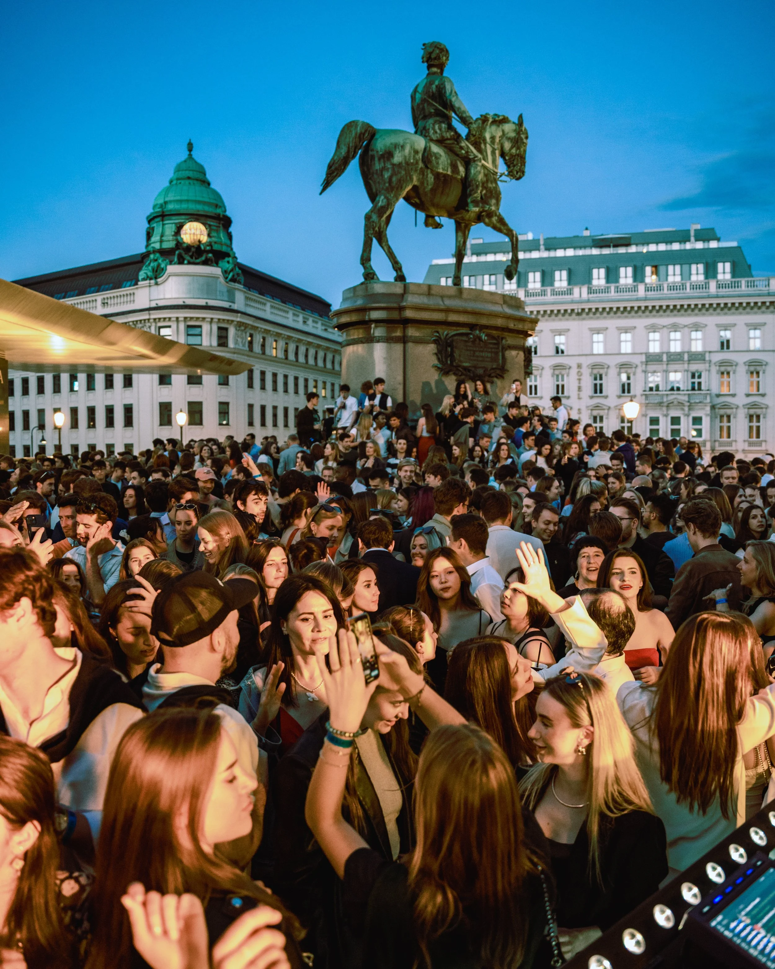 Crowd of people at an outdoor event during evening with an equestrian statue and historic buildings in the background.
