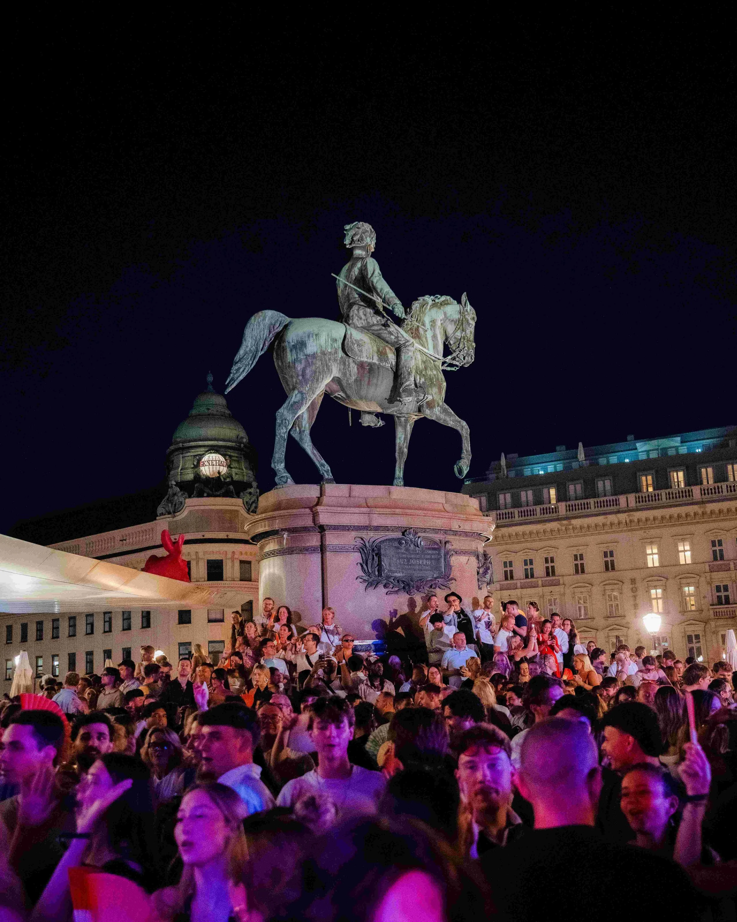 Crowd gathered at a public square during nighttime, with an illuminated equestrian statue of a man on a horse in the background, and surrounding historic buildings.