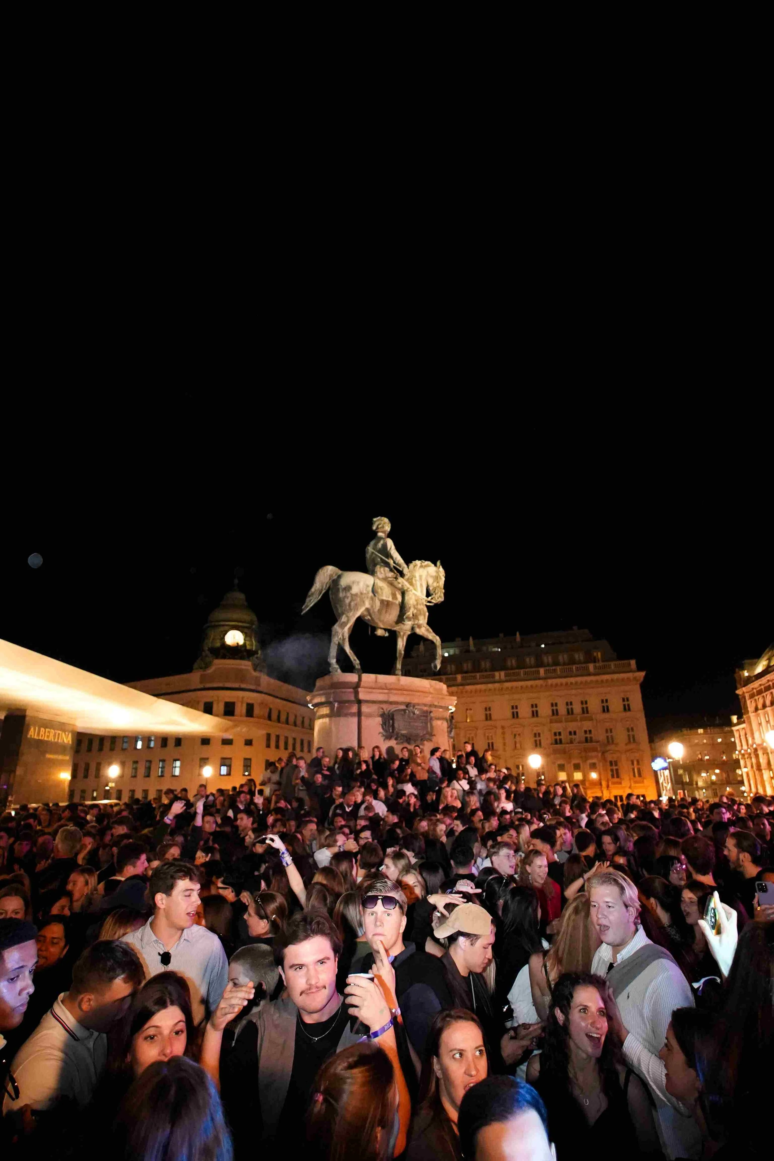 Nighttime scene at a crowded square with a large equestrian statue of a man in military attire on a horse, illuminated buildings, and a crowd of people socializing and taking photos.