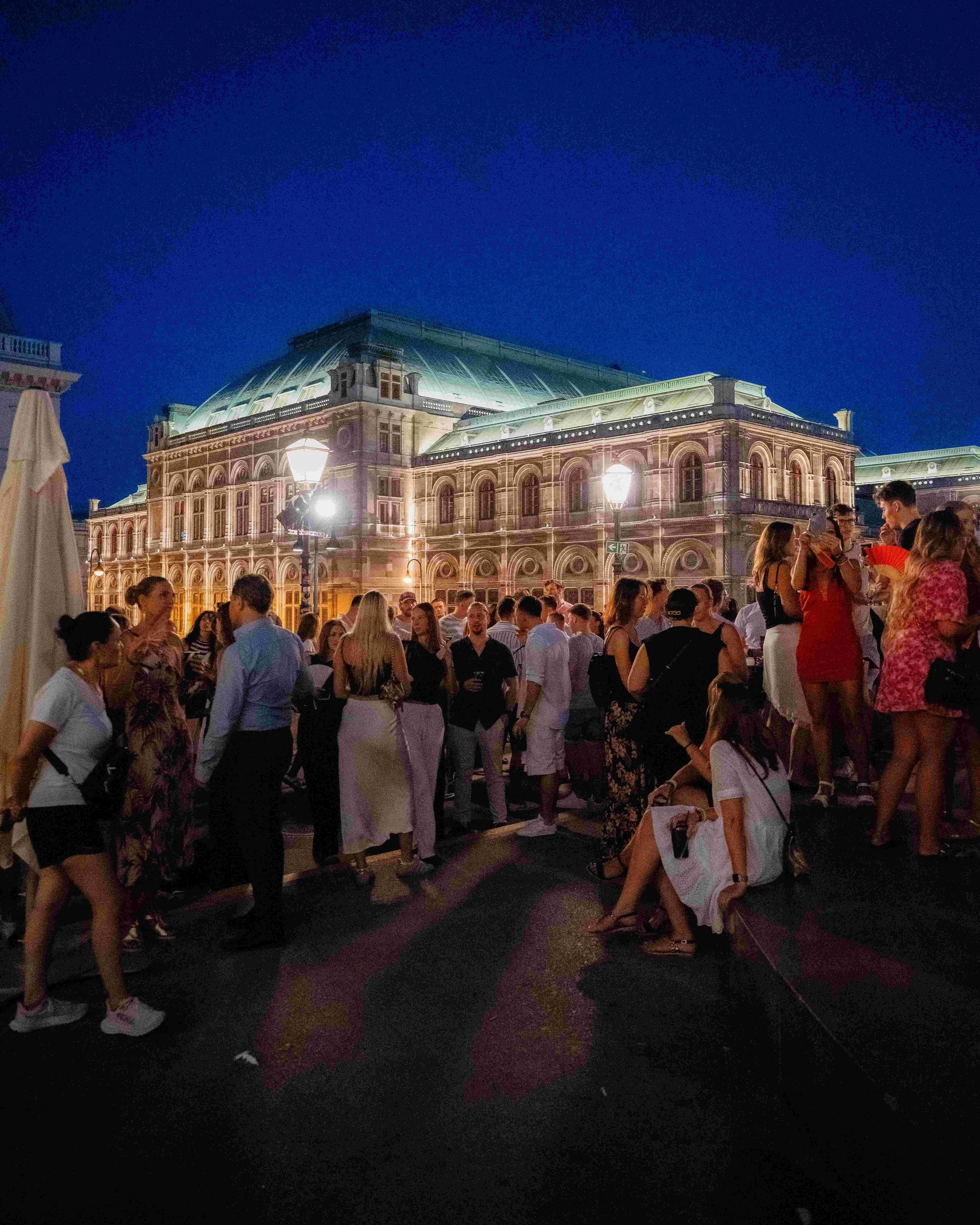 Night view of a grand historic building illuminated by warm lighting, with a large crowd of people gathered in front, some standing and some sitting, under street lamps and a clear dark blue sky.