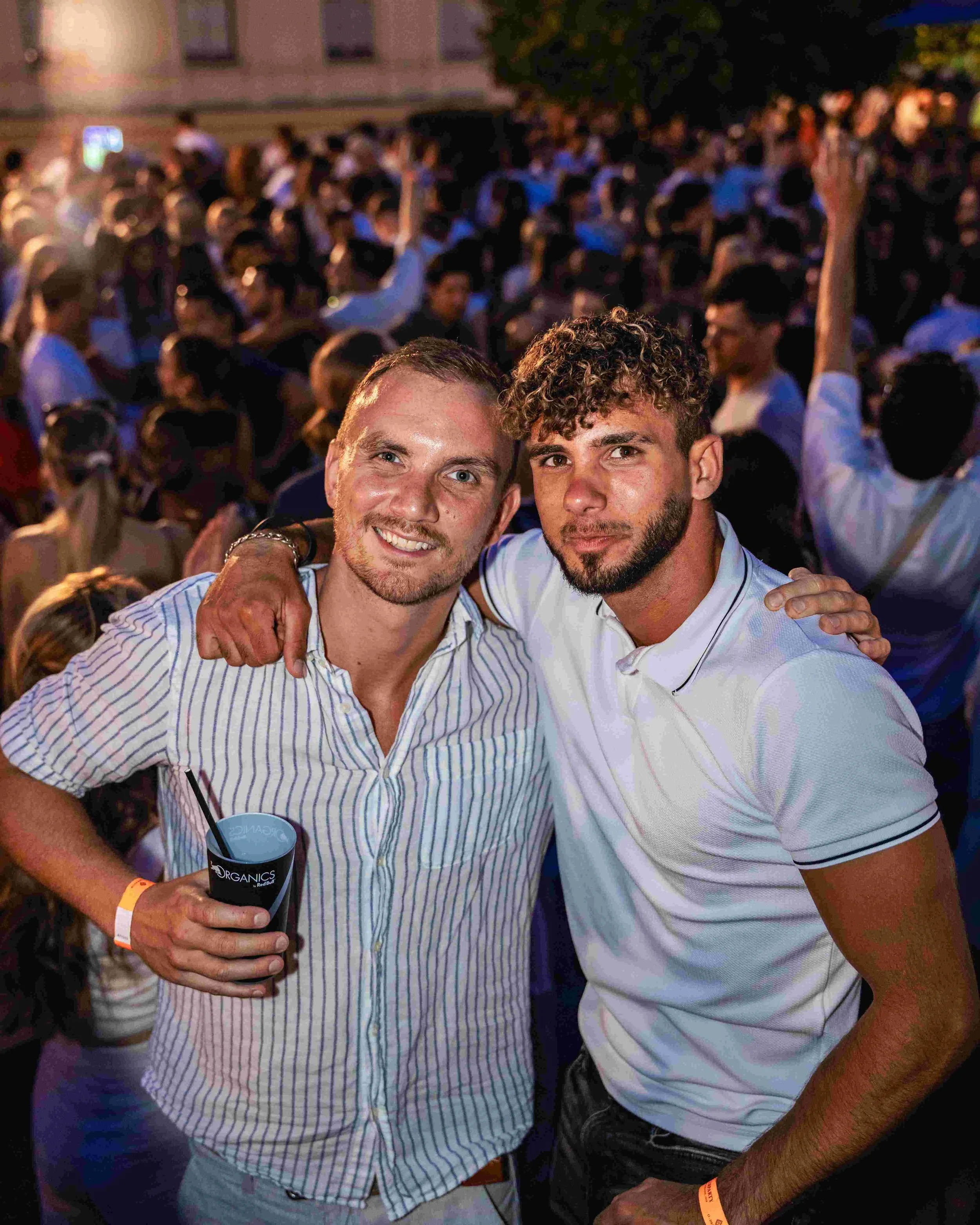 Two young men smiling at a lively outdoor event, one holding a drink, both wearing casual shirts, with many people in the blurred background.