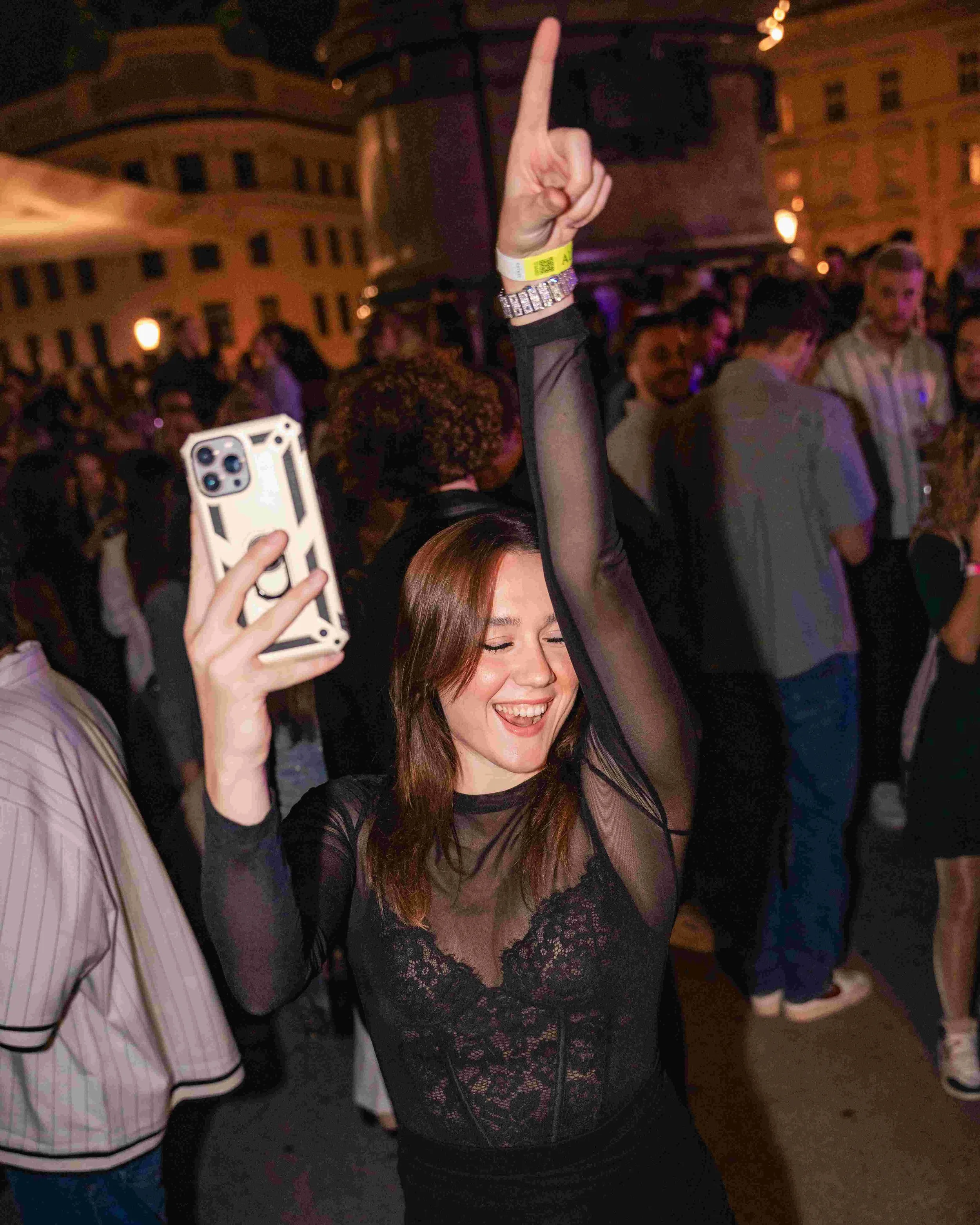 A woman with reddish-brown hair, smiling with eyes closed, wearing a black lace top, is raising her right hand with her index finger pointing upwards at a lively event with people in the background. A person next to her is holding a smartphone.