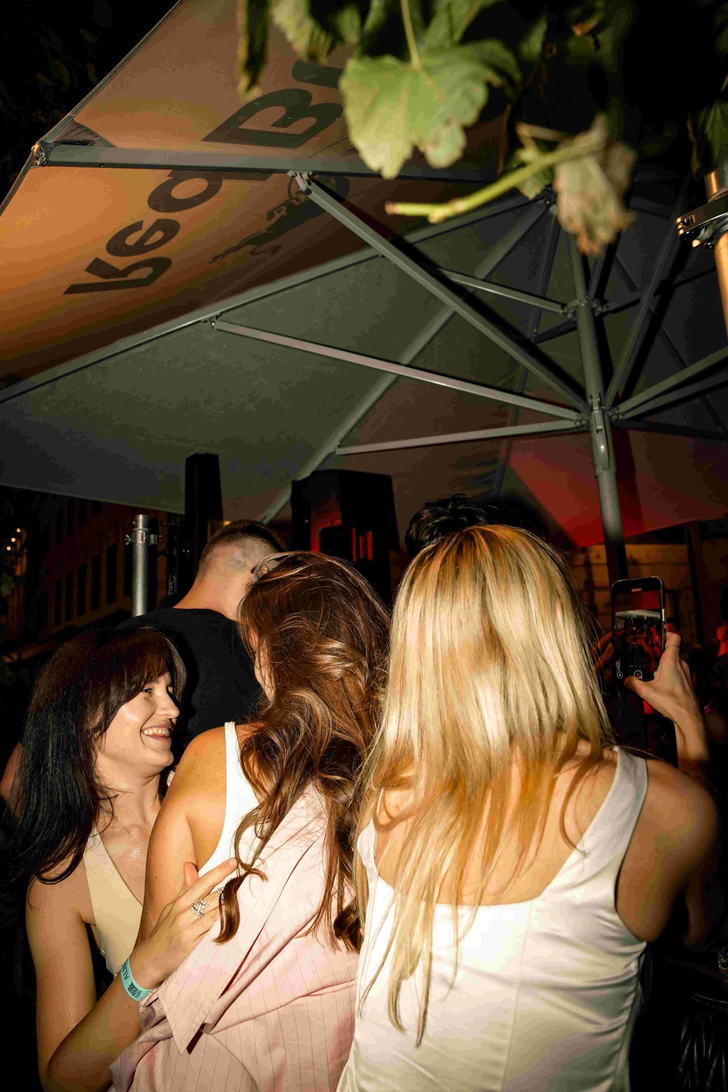 A group of women smiling and embracing each other at a social event, with a large umbrella and a person taking a photo in the background.