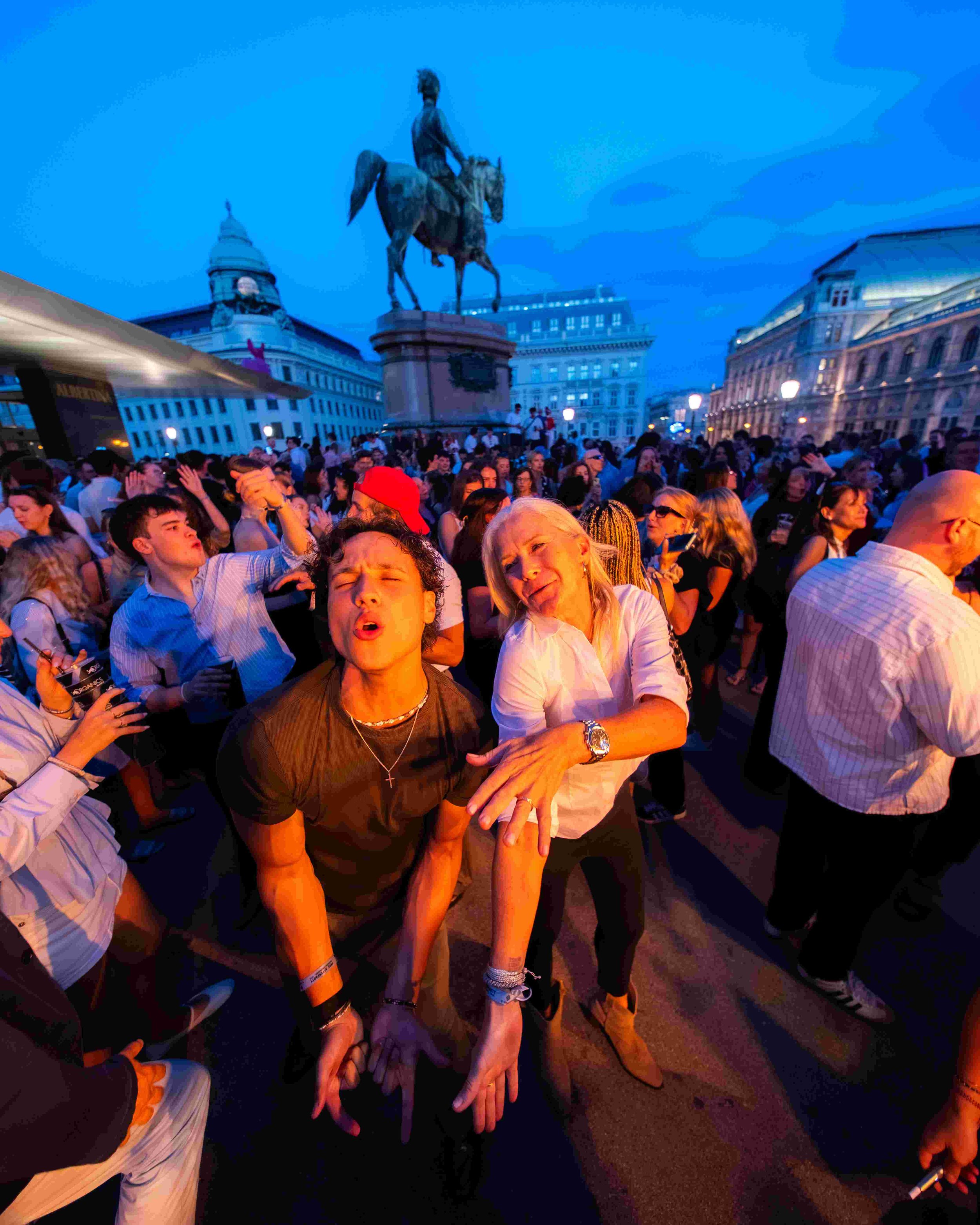 Crowd of people gathered at an outdoor event during twilight, with a statue of a mounted figure on a horse in the background and historic buildings with bright lights surrounding the scene.