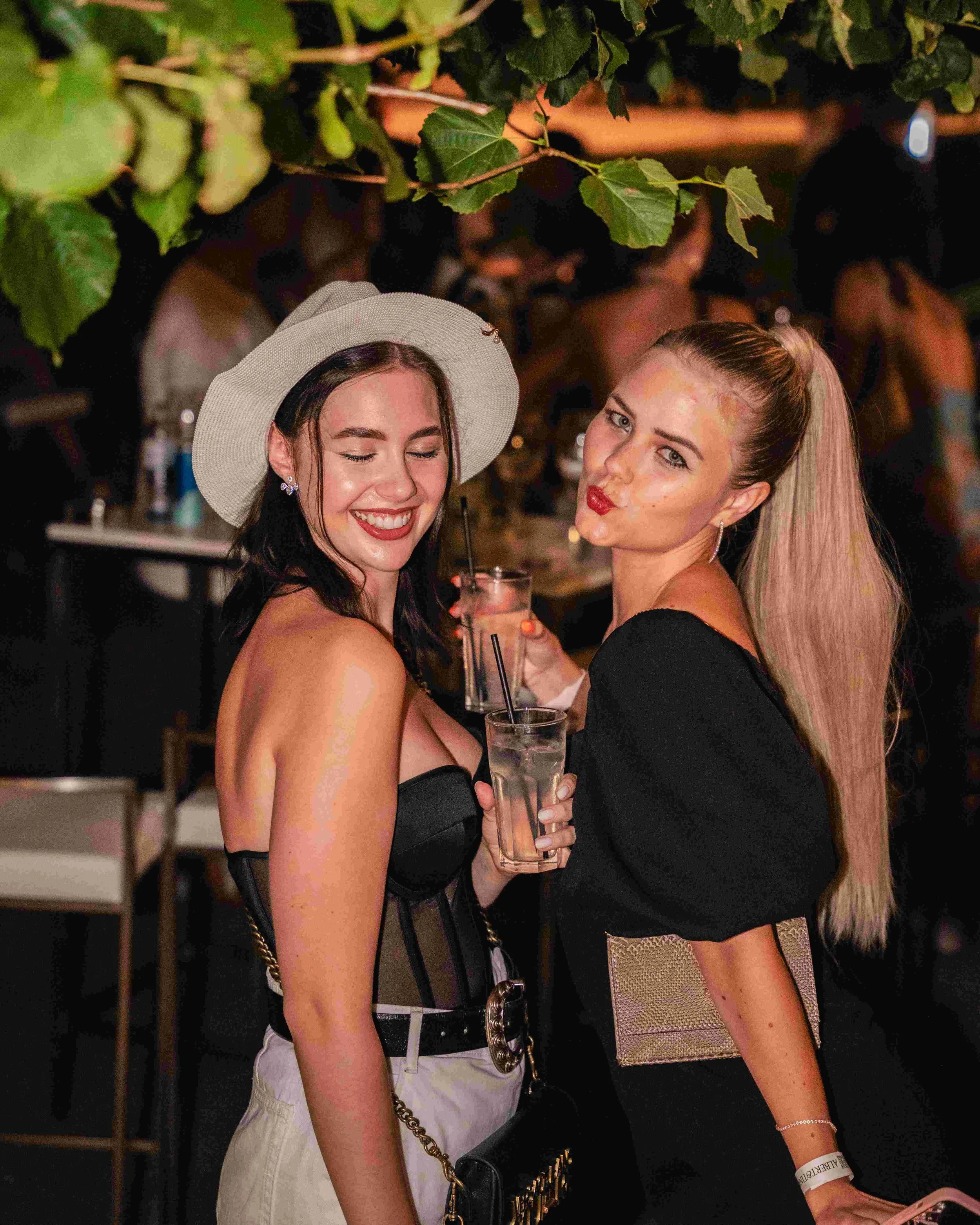Two women at a social event, smiling and holding drinks, dressed in stylish outfits. One woman wears a wide-brimmed hat and white pants; the other has long blonde hair and a black dress.