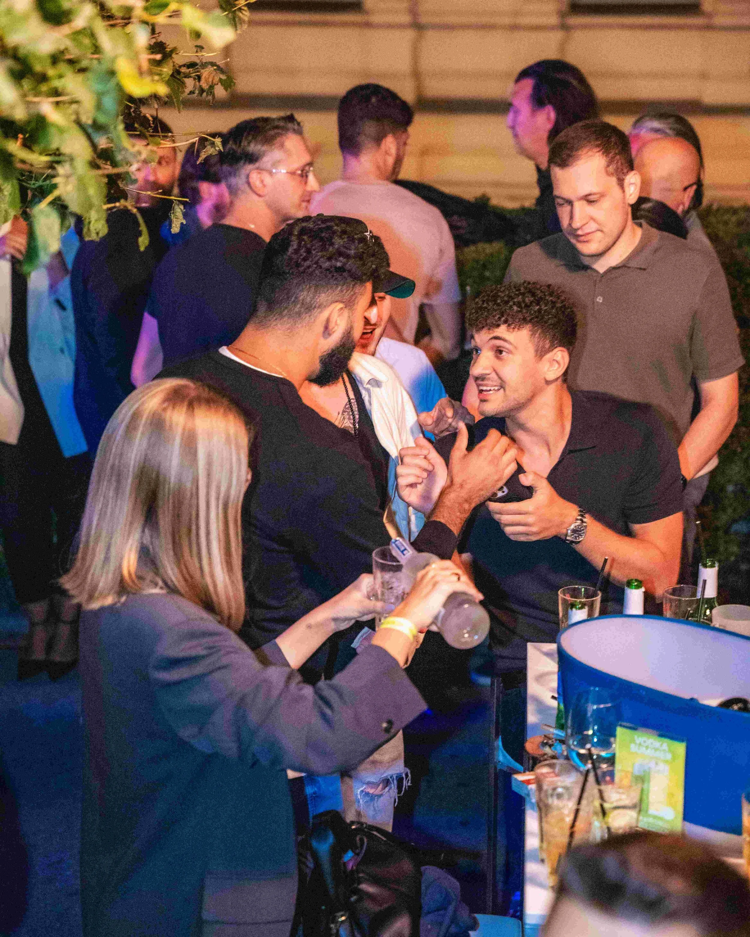 Group of people socializing at an outdoor event, with some holding drinks and engaging in conversation at a table with snacks and drinks in a dimly lit setting.