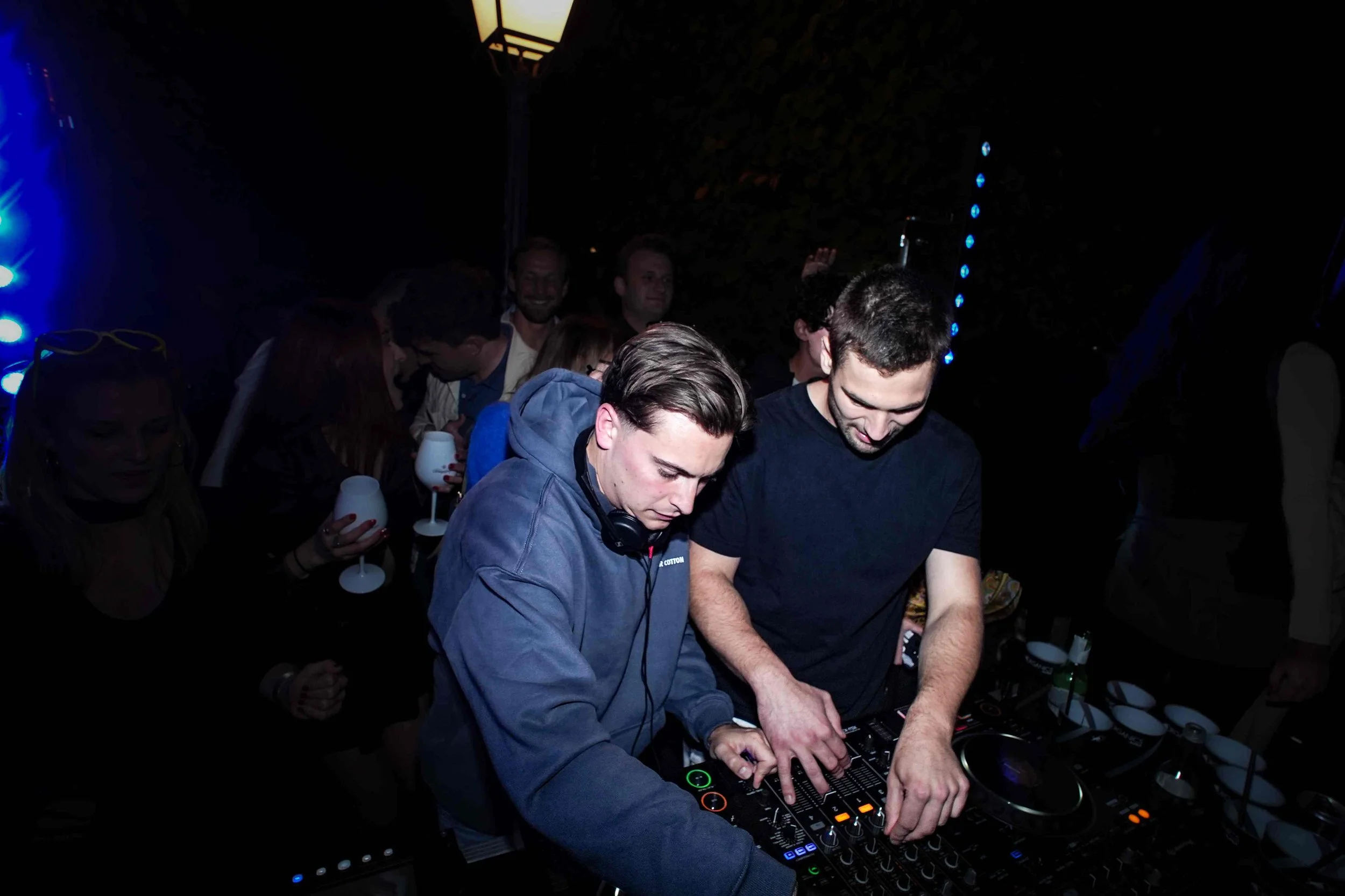 Two male DJs are performing at a nightclub, surrounded by a crowd of people. The DJs are focused on their equipment, with one wearing headphones. The scene is dark with blue and purple lighting.