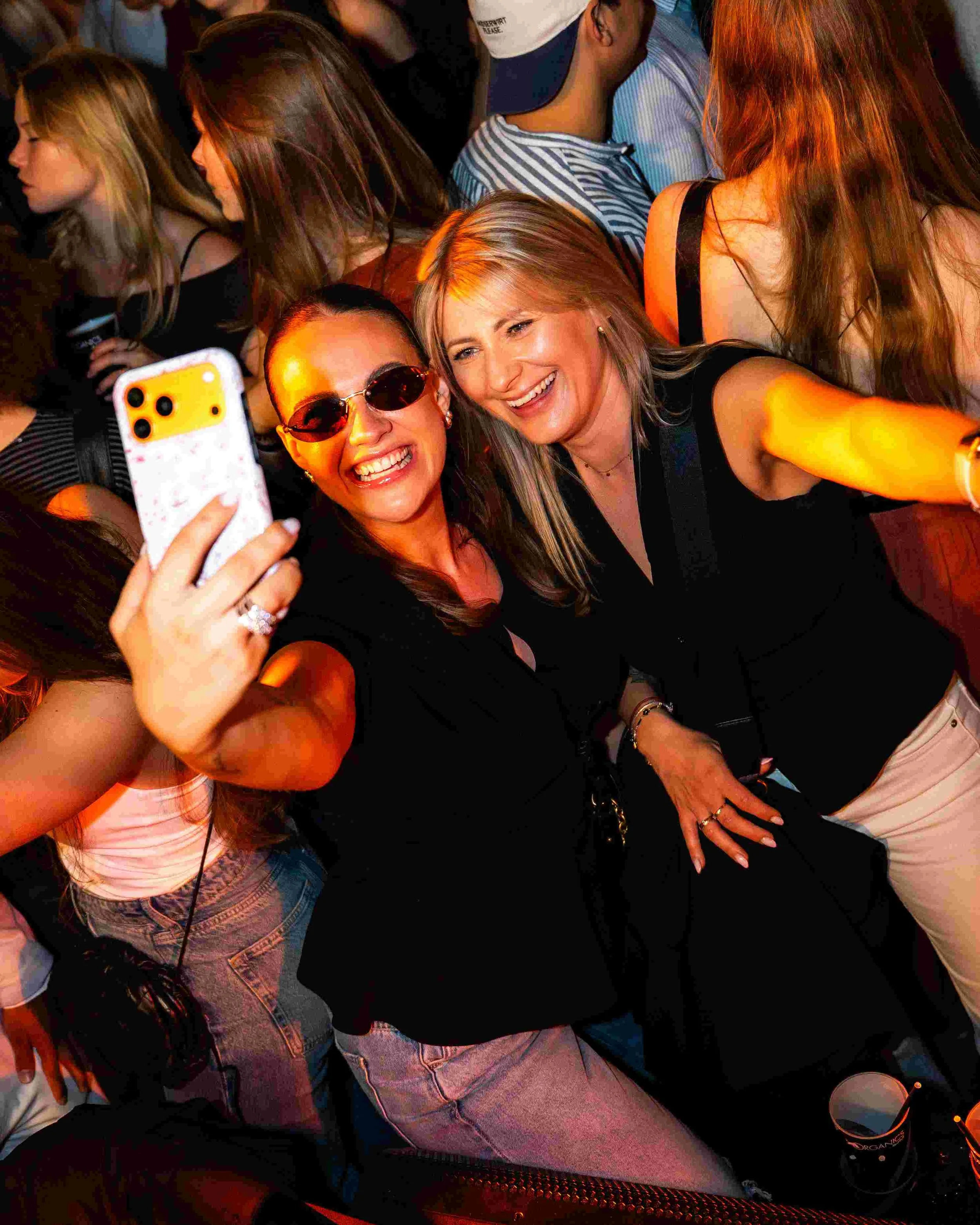 A group of people taking a selfie at a crowded party or event. Two women are prominently in the foreground, smiling and posing for the camera. One woman is wearing sunglasses and jewelry, while the other has blonde hair and is wearing a black top. Mu