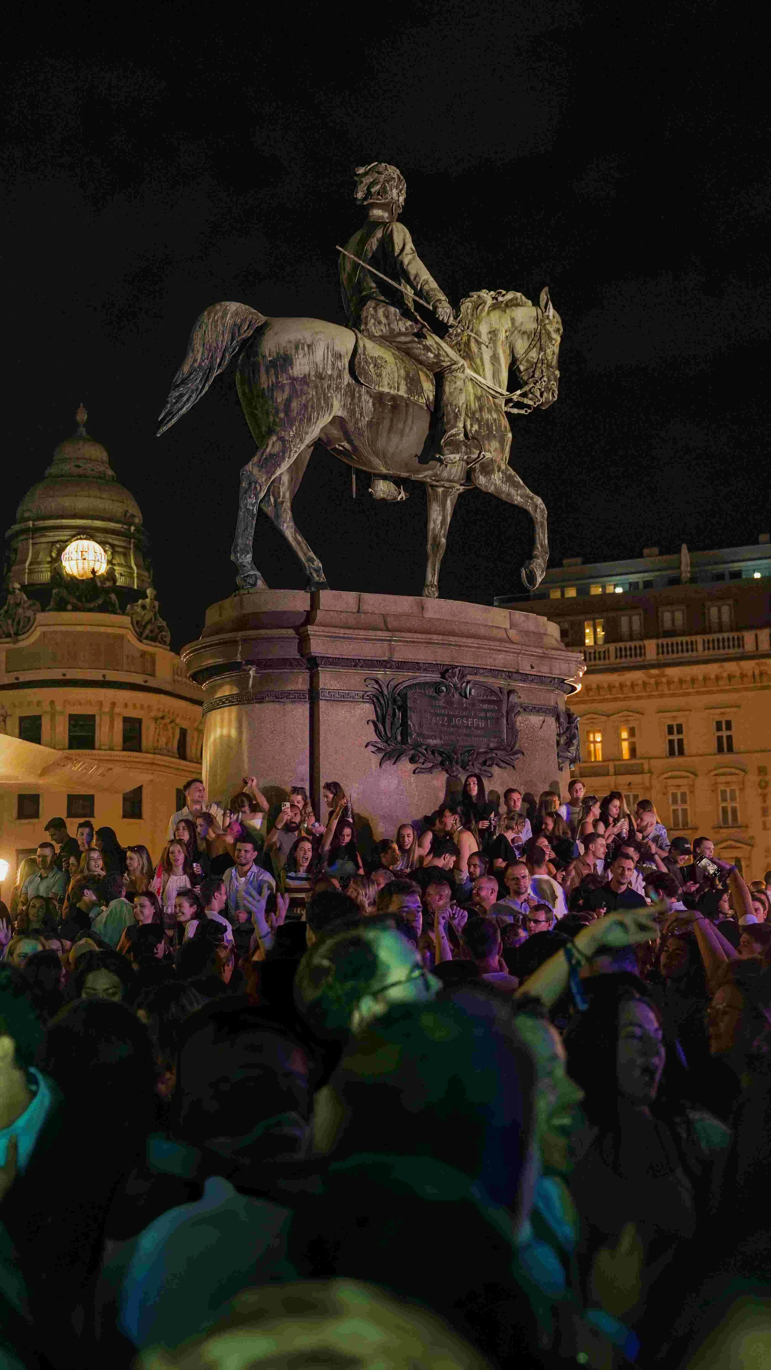Nighttime view of an equestrian statue of a man on horseback on a large pedestal, surrounded by a densely crowded group of people, with historic buildings illuminated in the background.