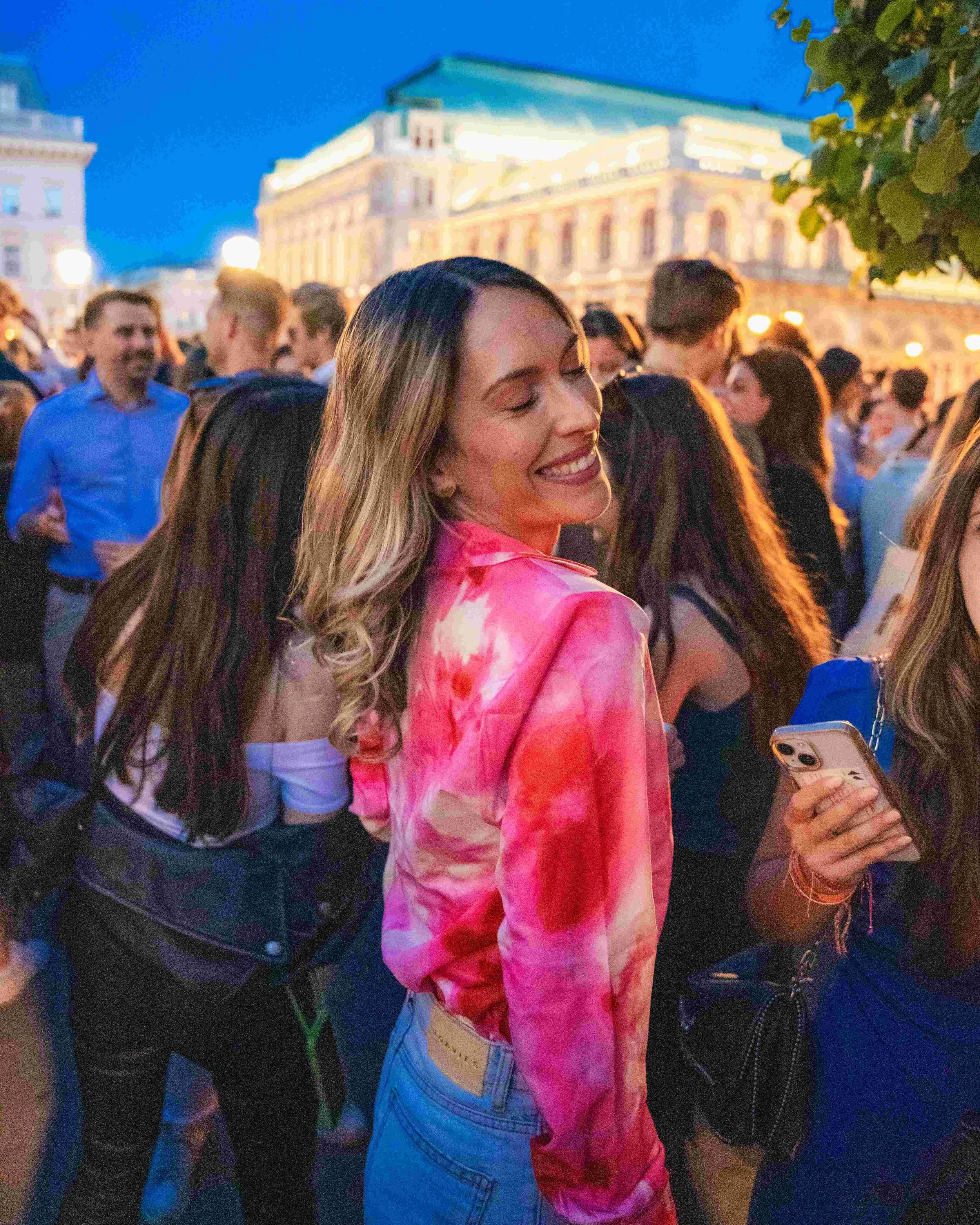 A young woman with long blonde hair, smiling with eyes closed, wearing a pink tie-dye shirt, standing in a crowd of people outdoors at night, with historic illuminated buildings in the background.