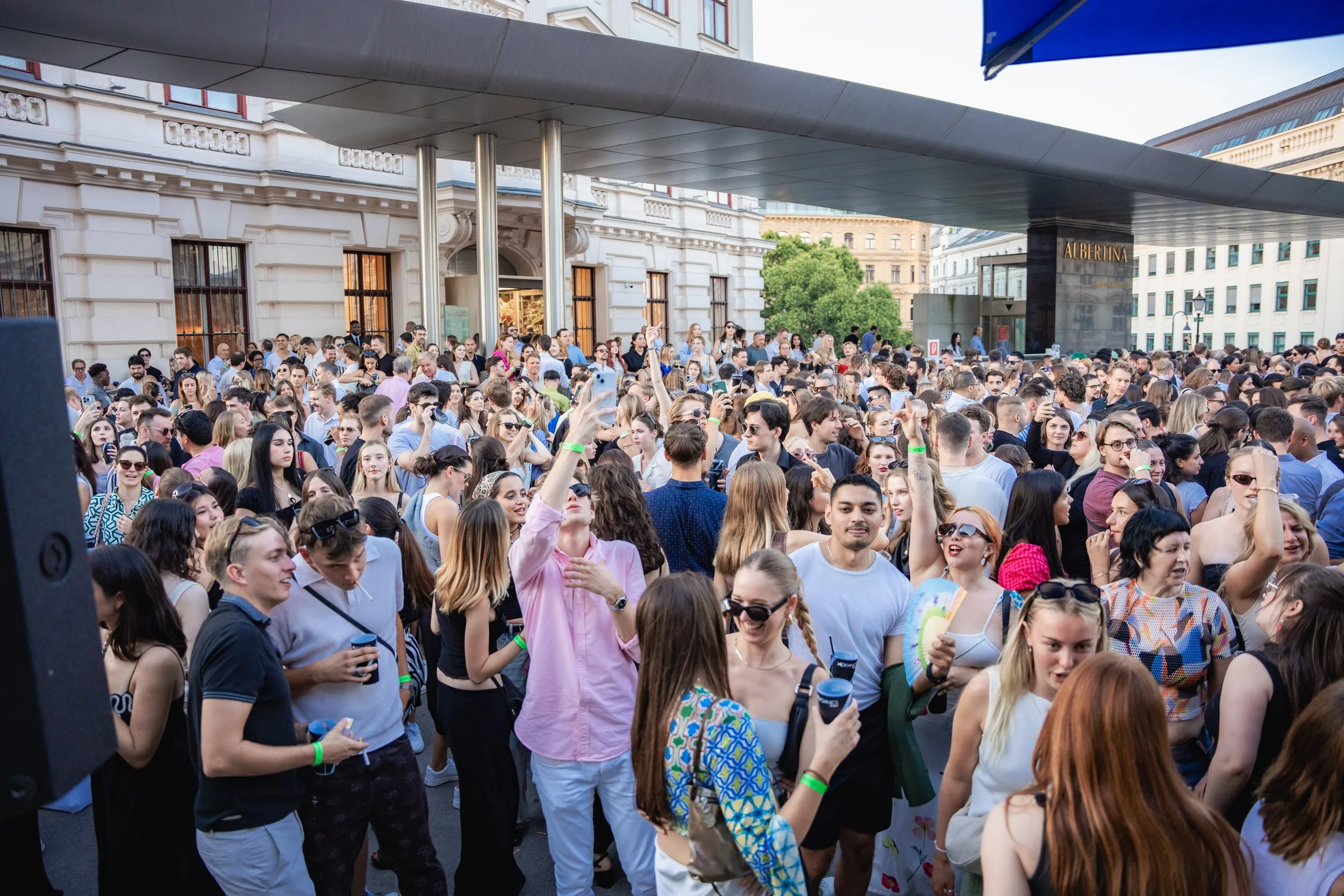 A large crowd of young people gathered outdoors in front of a historic building with white stone walls and large windows, some holding drinks and taking photos.