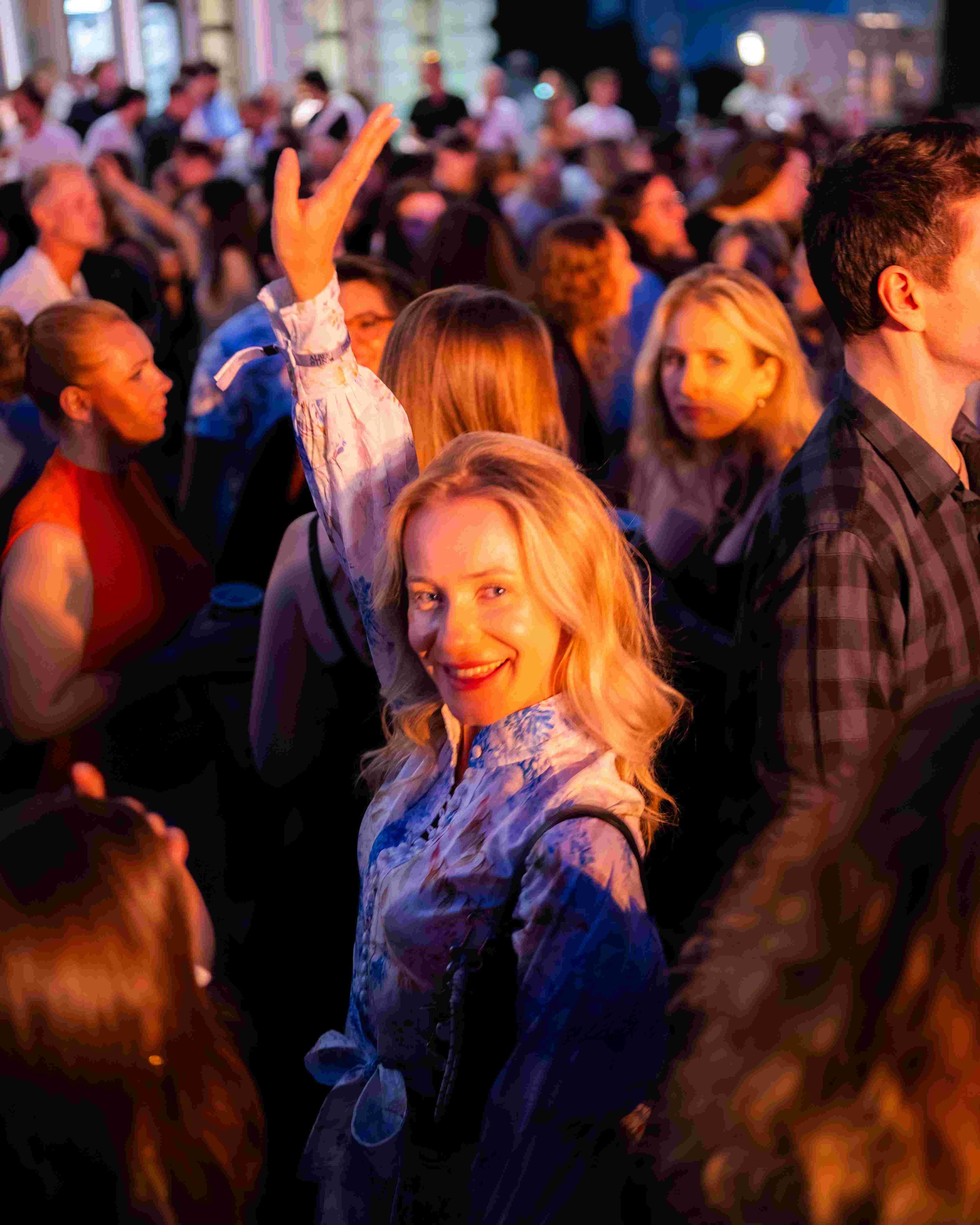 A diverse group of people attending an indoor event, some raising hands, with ambient colorful lighting and a lively atmosphere.