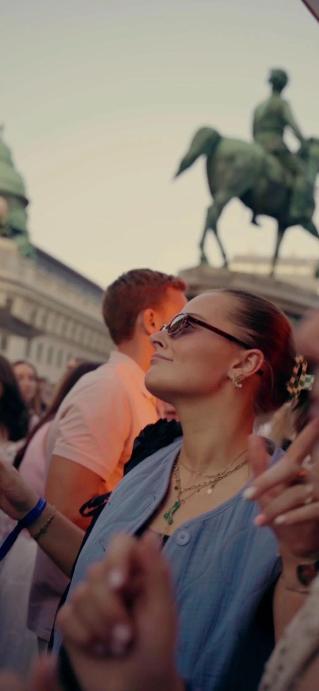 A woman wearing sunglasses and jewelry smiling and enjoying a crowd during sunset with a statue on horseback visible in the background.