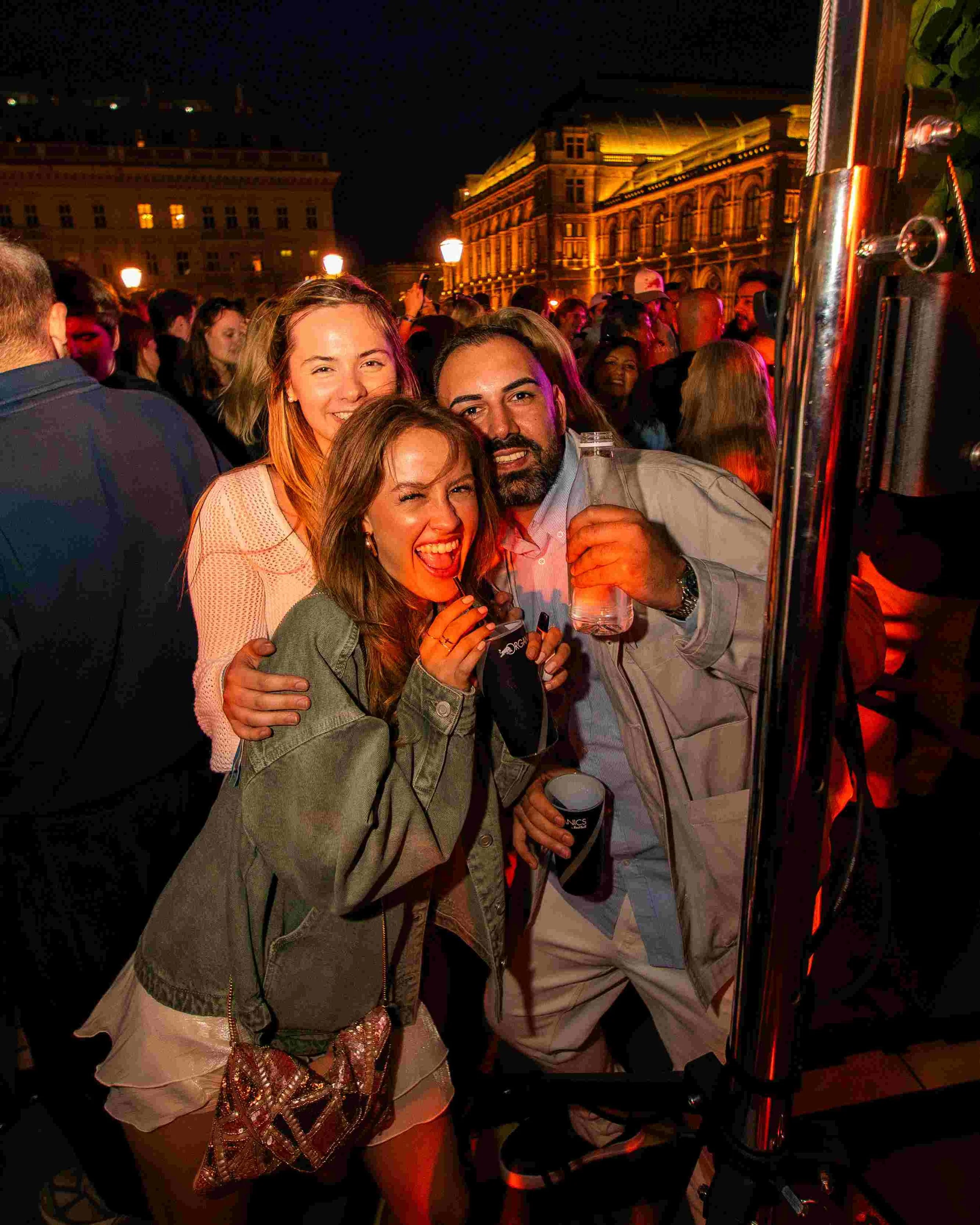 A group of four people, three women and one man, smiling and posing for a photo at a nighttime outdoor event. They are close together, with the women on the left and the man on the right. The background shows a crowd of people and illuminated histori