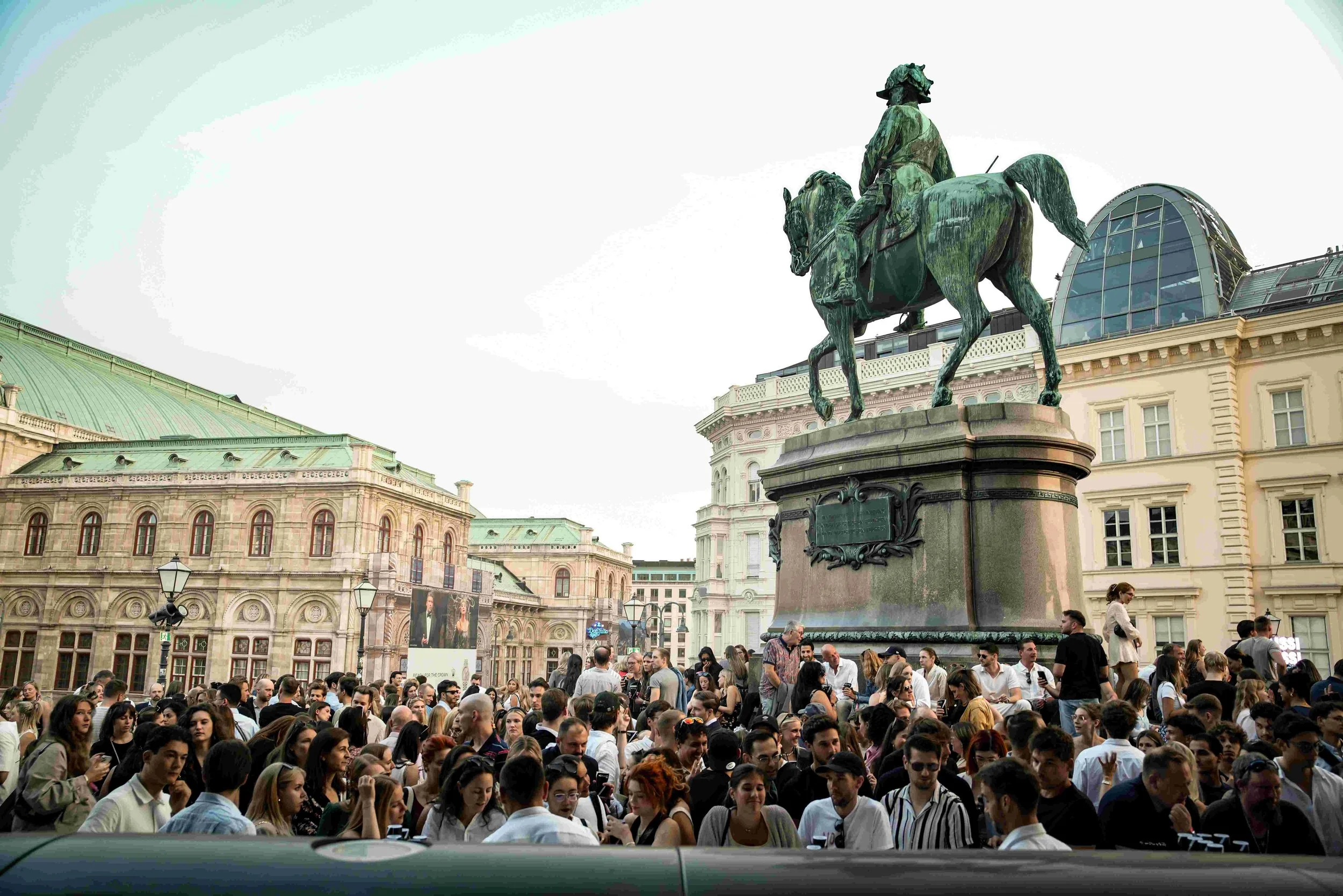 A large crowd of people gathered in a historic square with an equestrian statue of a man in military attire on horseback. The background features ornate historic buildings and a modern glass-domed structure.