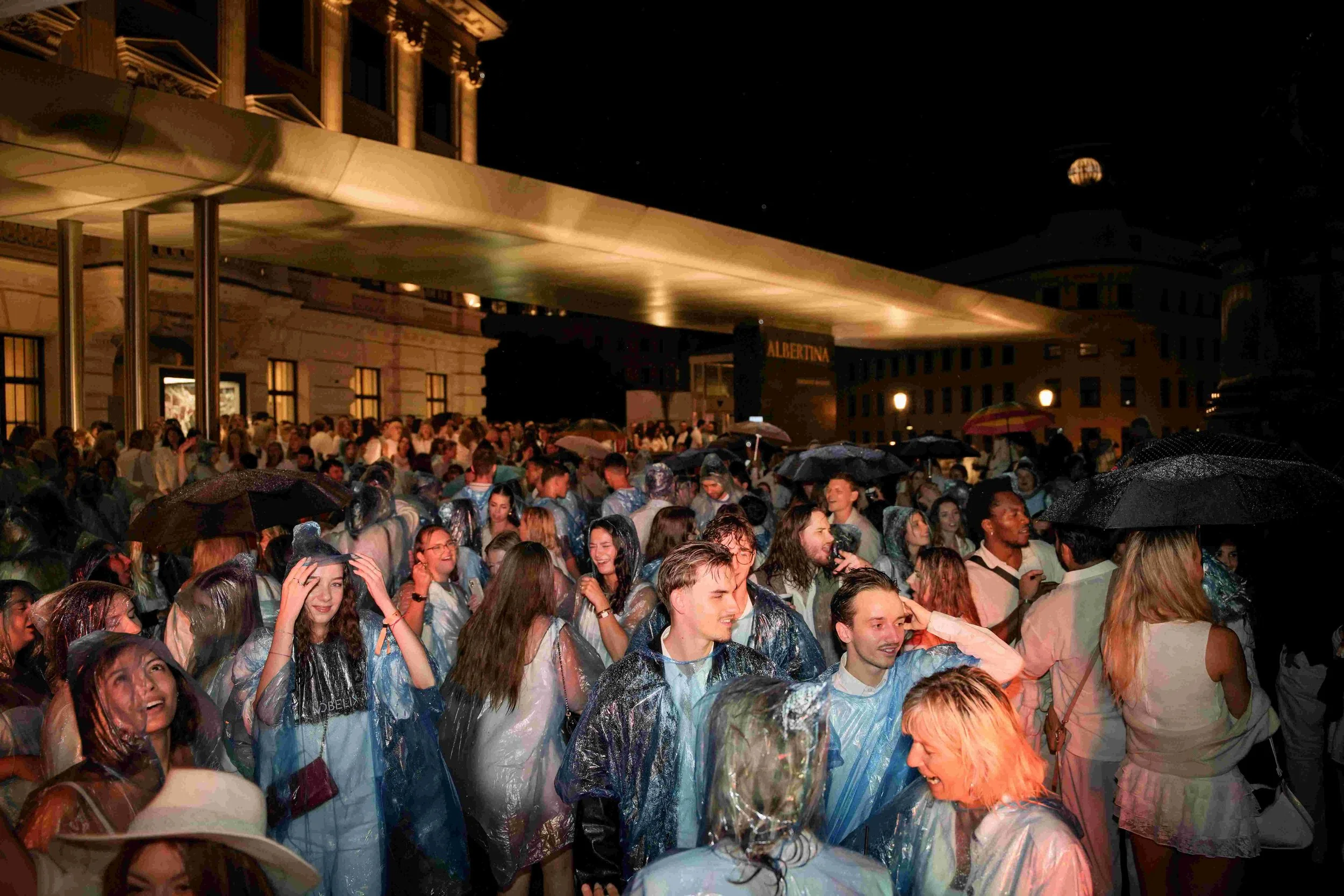 A large crowd of people gathered outdoors at night, many wearing rain ponchos, with some umbrellas visible, in front of a building with a balcony and a sign that reads 'ALBERTINA' under a dark sky.