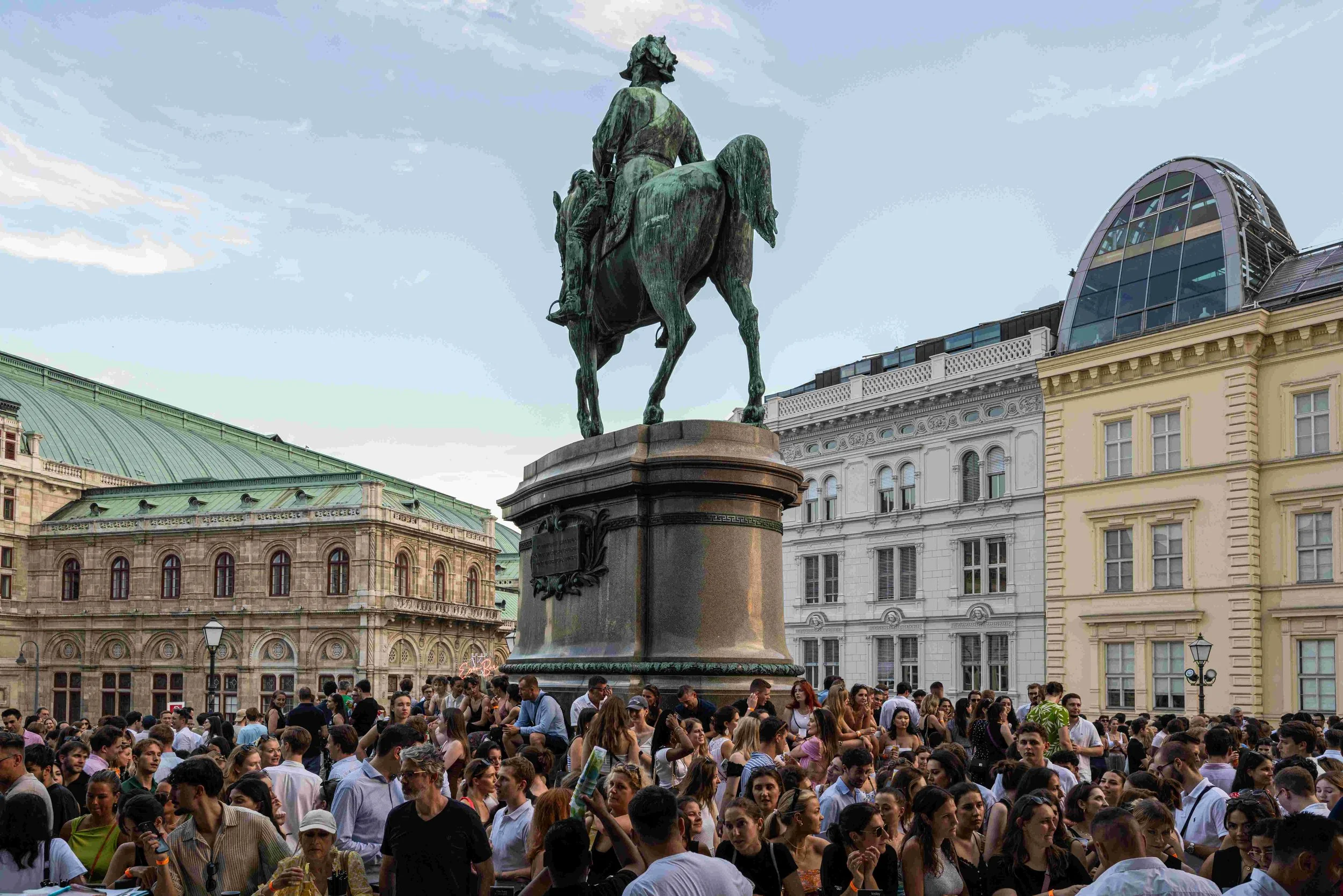 Crowd of people gathered around a statue of a man on horseback in an urban square with historic European-style buildings.