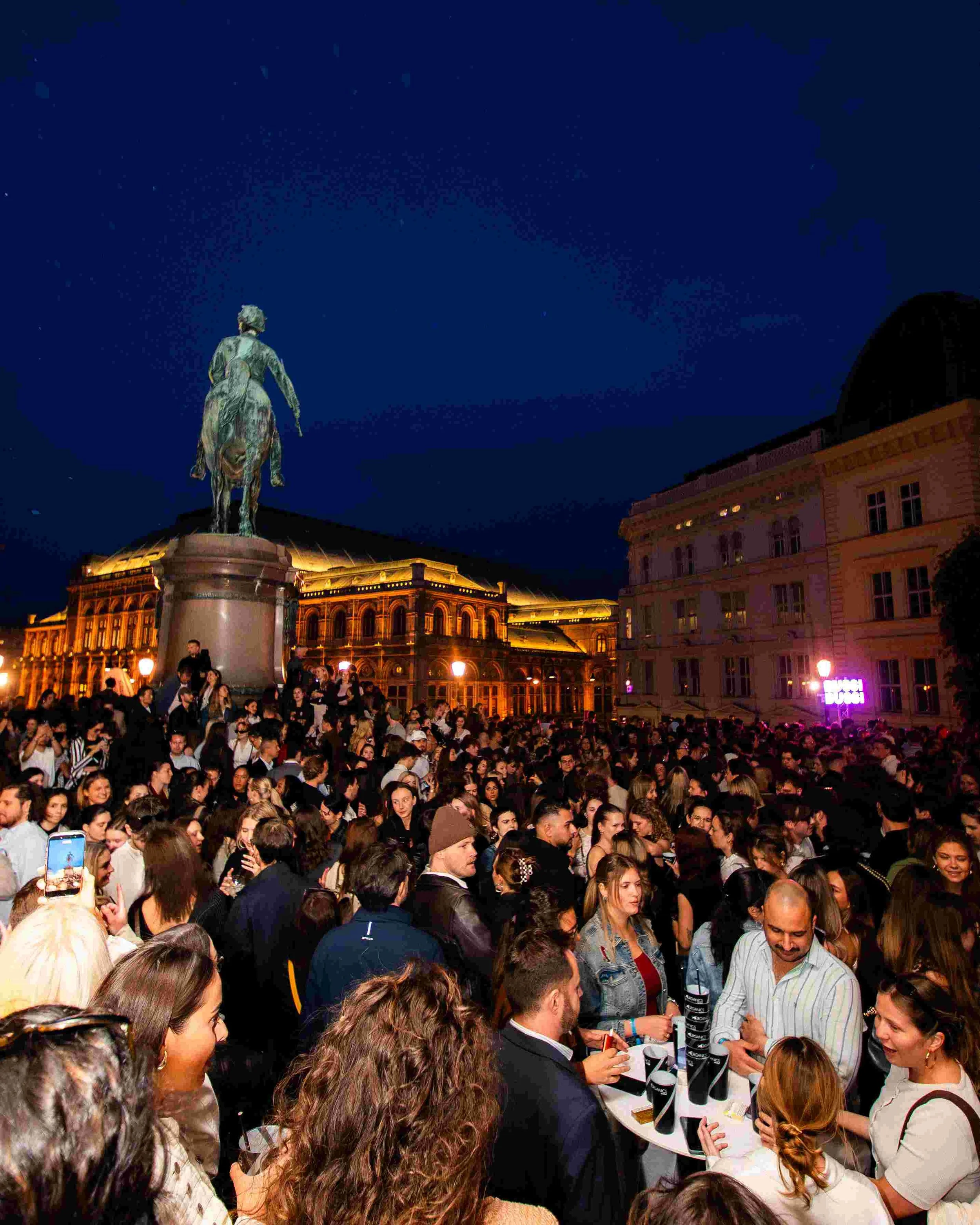 A crowd of people gathered in a city square at night with a statue of a man on a horse in the background, illuminated by streetlights.