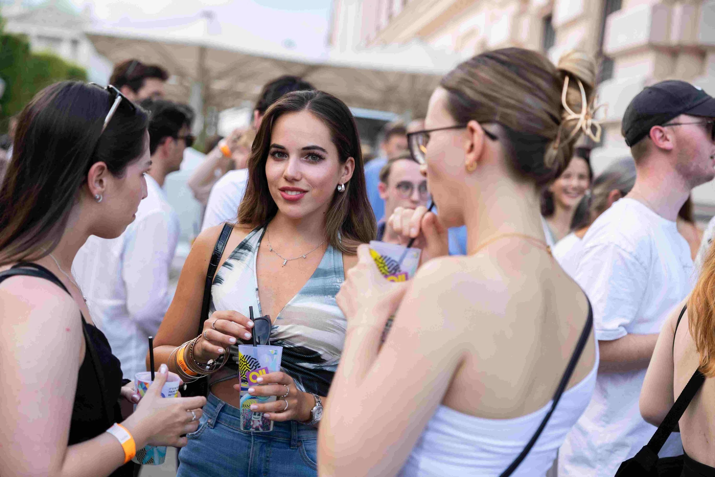 Group of young women socializing outdoors at a crowded event, holding drinks and engaging in conversation.