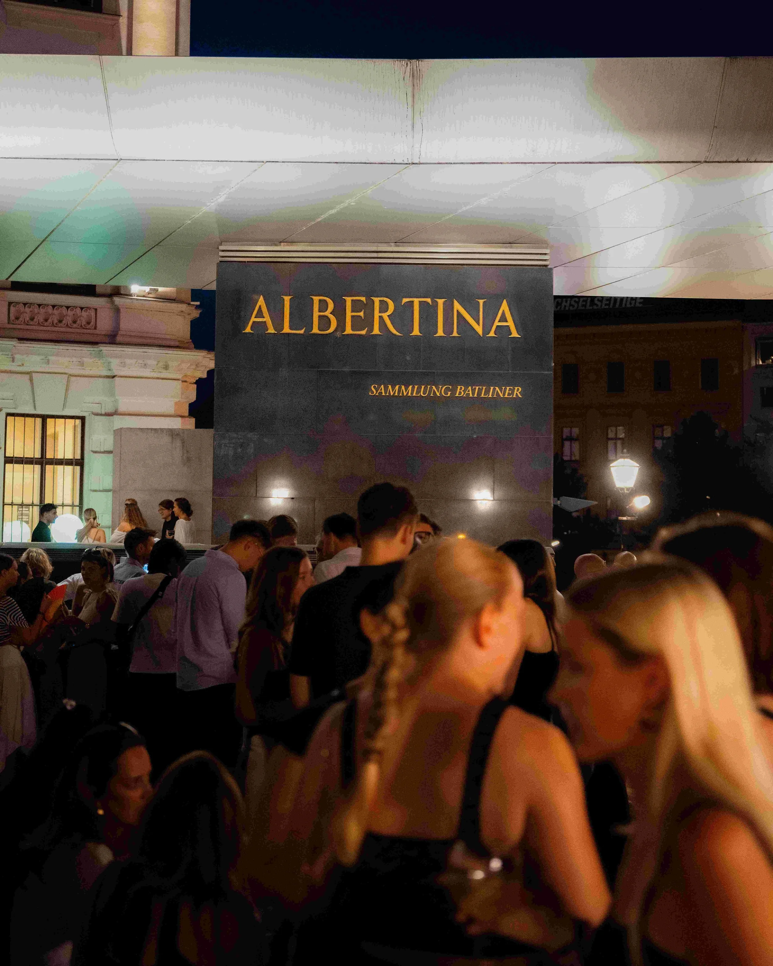 Nighttime scene outside a building with a sign that reads 'ALBERTINA' and 'SAMMLUNG BATLINER'. A group of people are gathered and socializing outside.