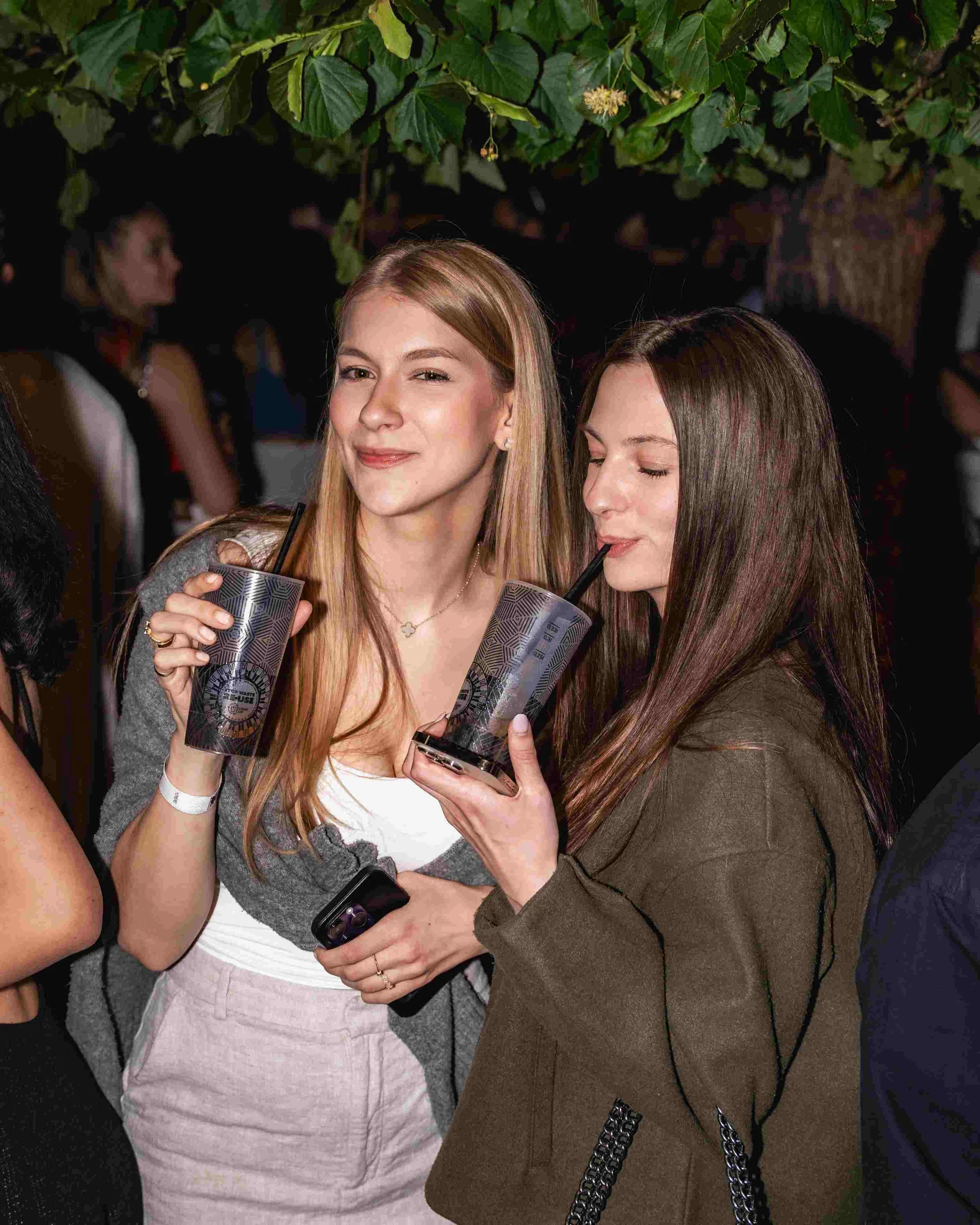 Two young women are holding drinks with black straws and smiling at each other at a social event, with one woman having long blonde hair and the other having long brown hair. A tree with large green leaves is visible above them.