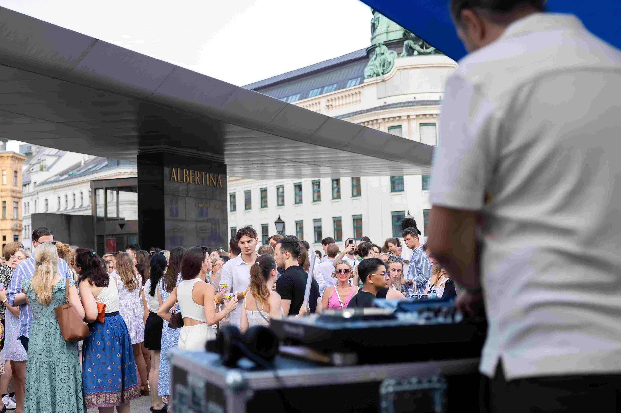 Crowd of people gathered outdoors near a building with a sign that reads 'ALBERTINA', with a person in the foreground using DJ equipment and a partial view of a person in a white shirt in the background.