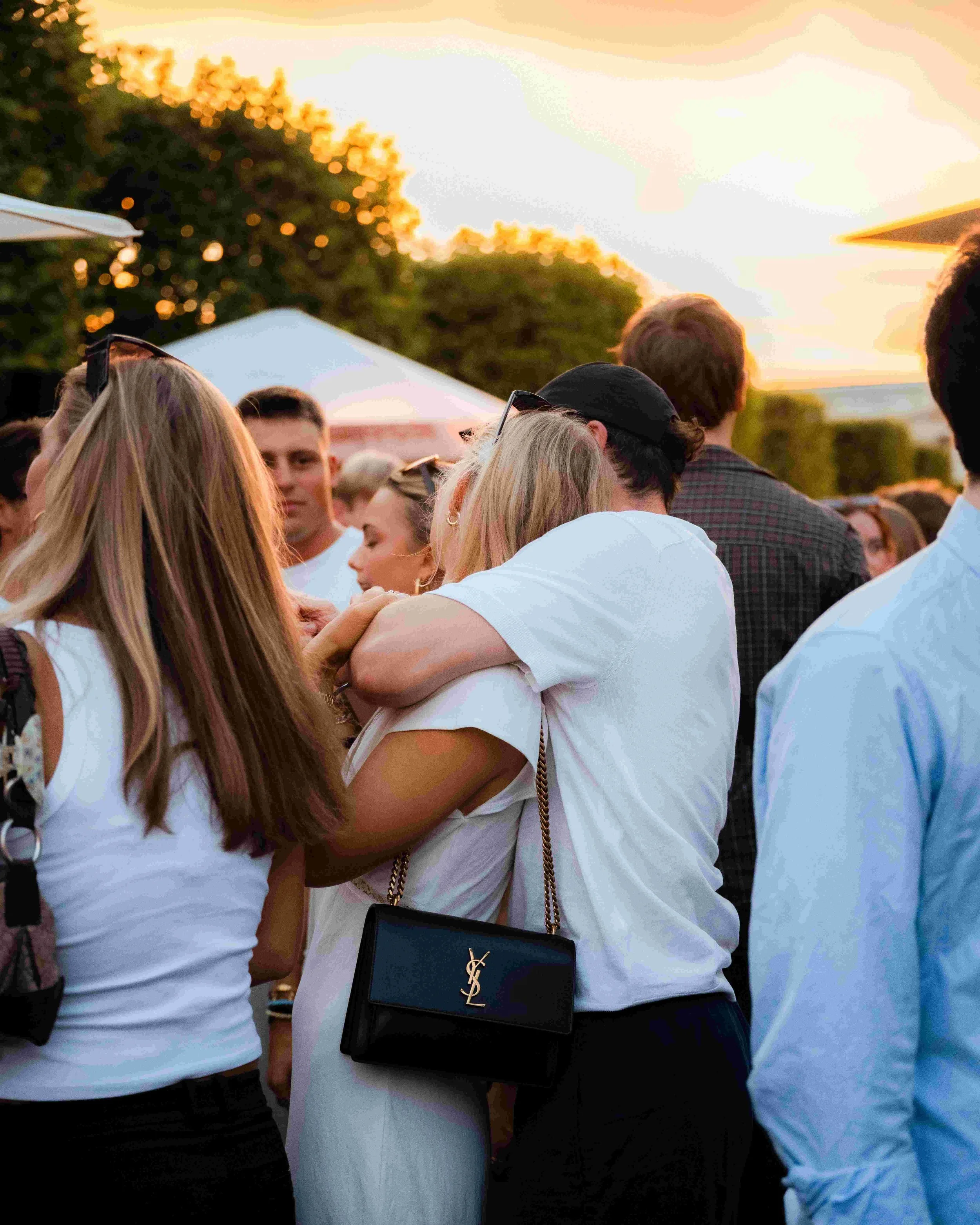 Group of people at an outdoor event during sunset, dressed casually, with many people facing away from the camera, some wearing sunglasses, and a person in the foreground with a Yves Saint Laurent bag.