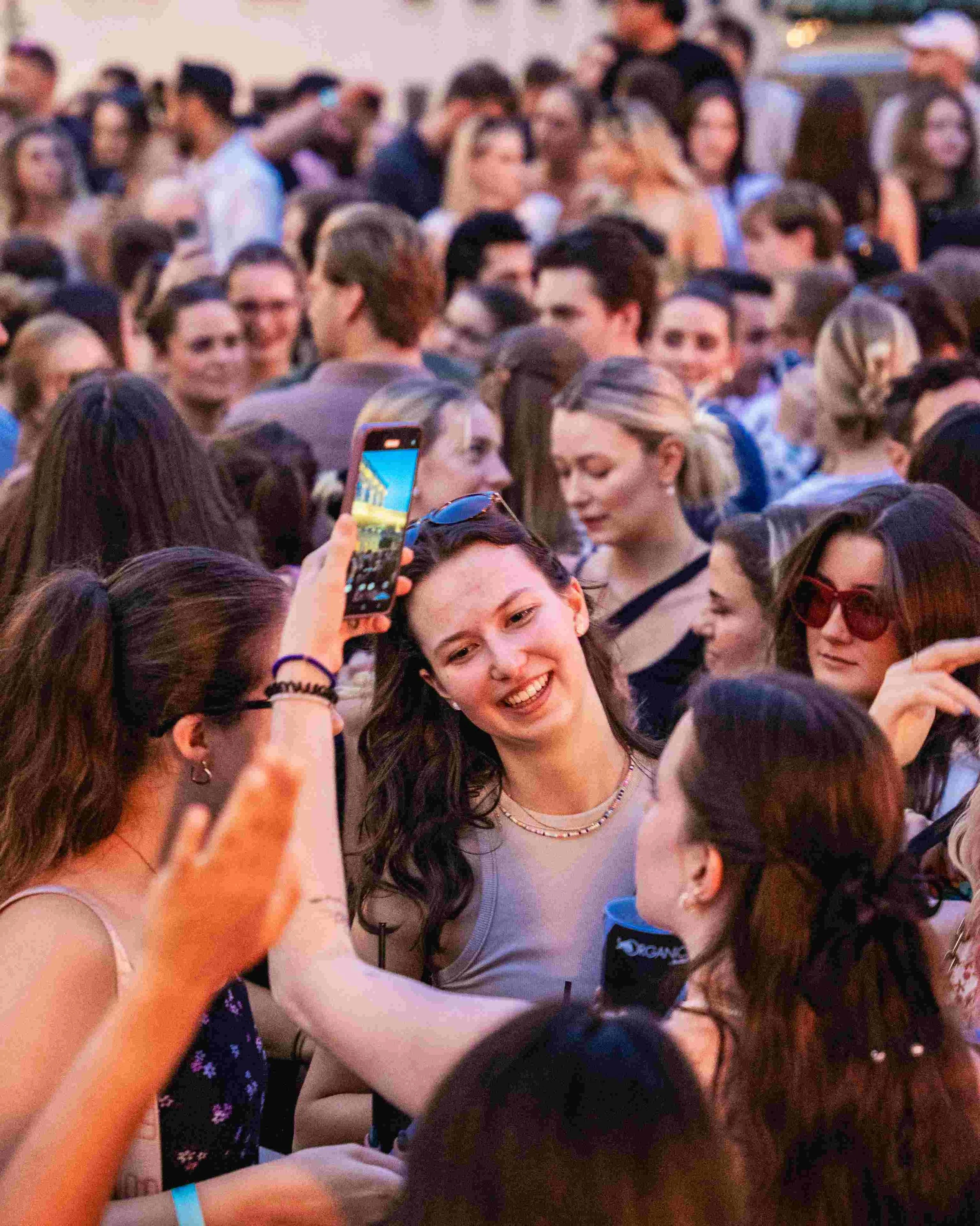 A large crowd of young people at an outdoor event, many smiling, engaging, and taking photos with their phones.