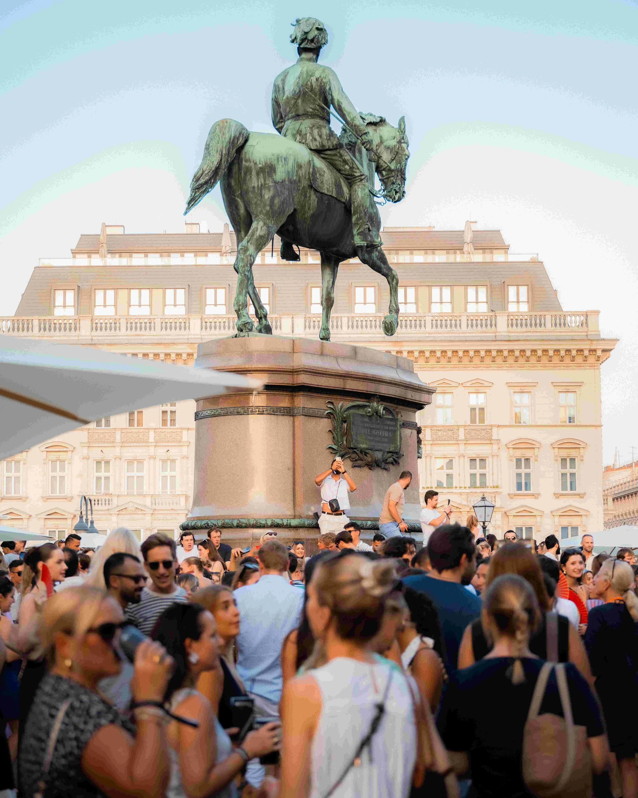 Crowd of people gathered in front of an equestrian statue in a city square, with historic buildings in the background.