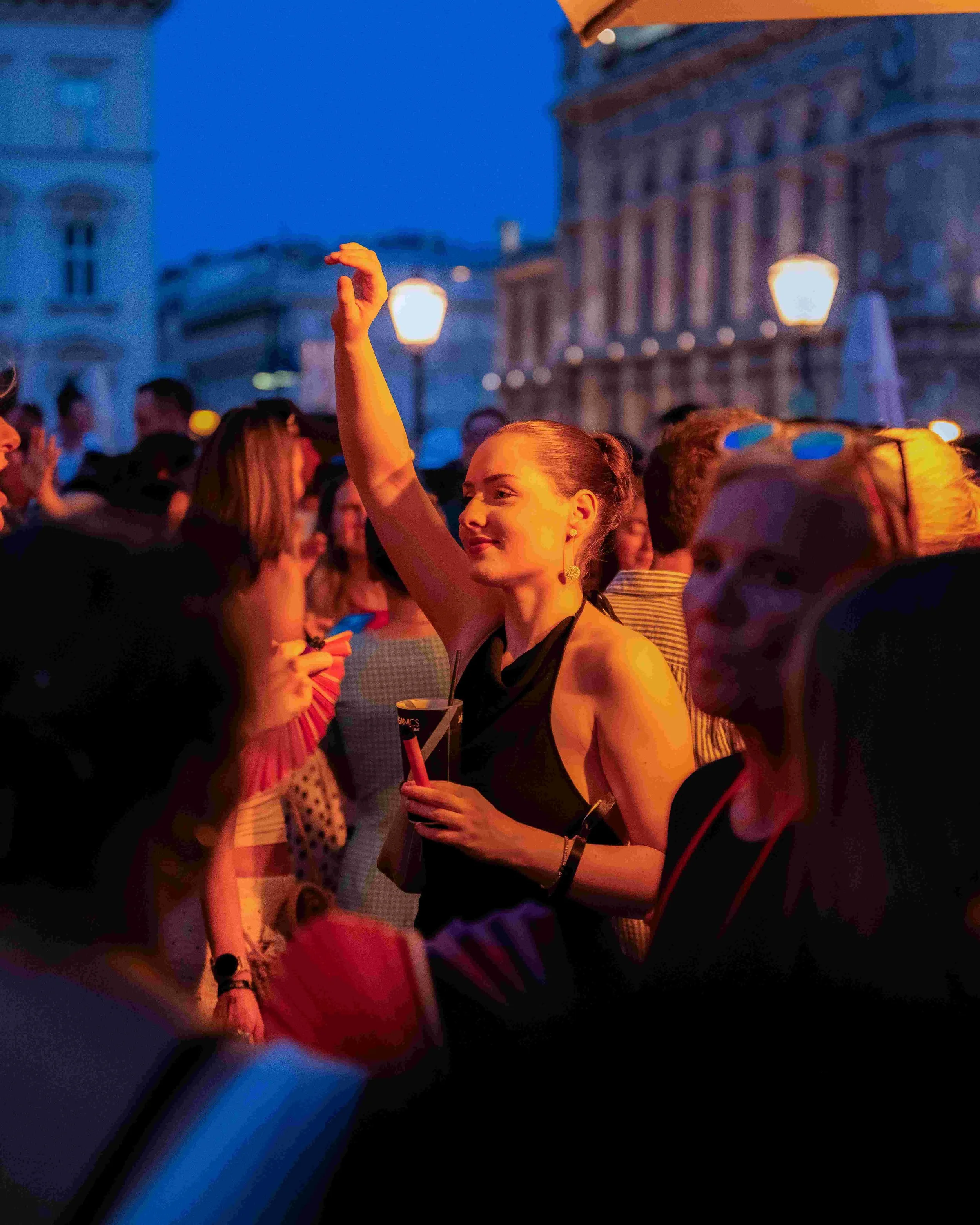 Crowd of people at an outdoor event during evening, with a woman in the foreground smiling and raising her arm, surrounded by blurred city buildings and streetlights.