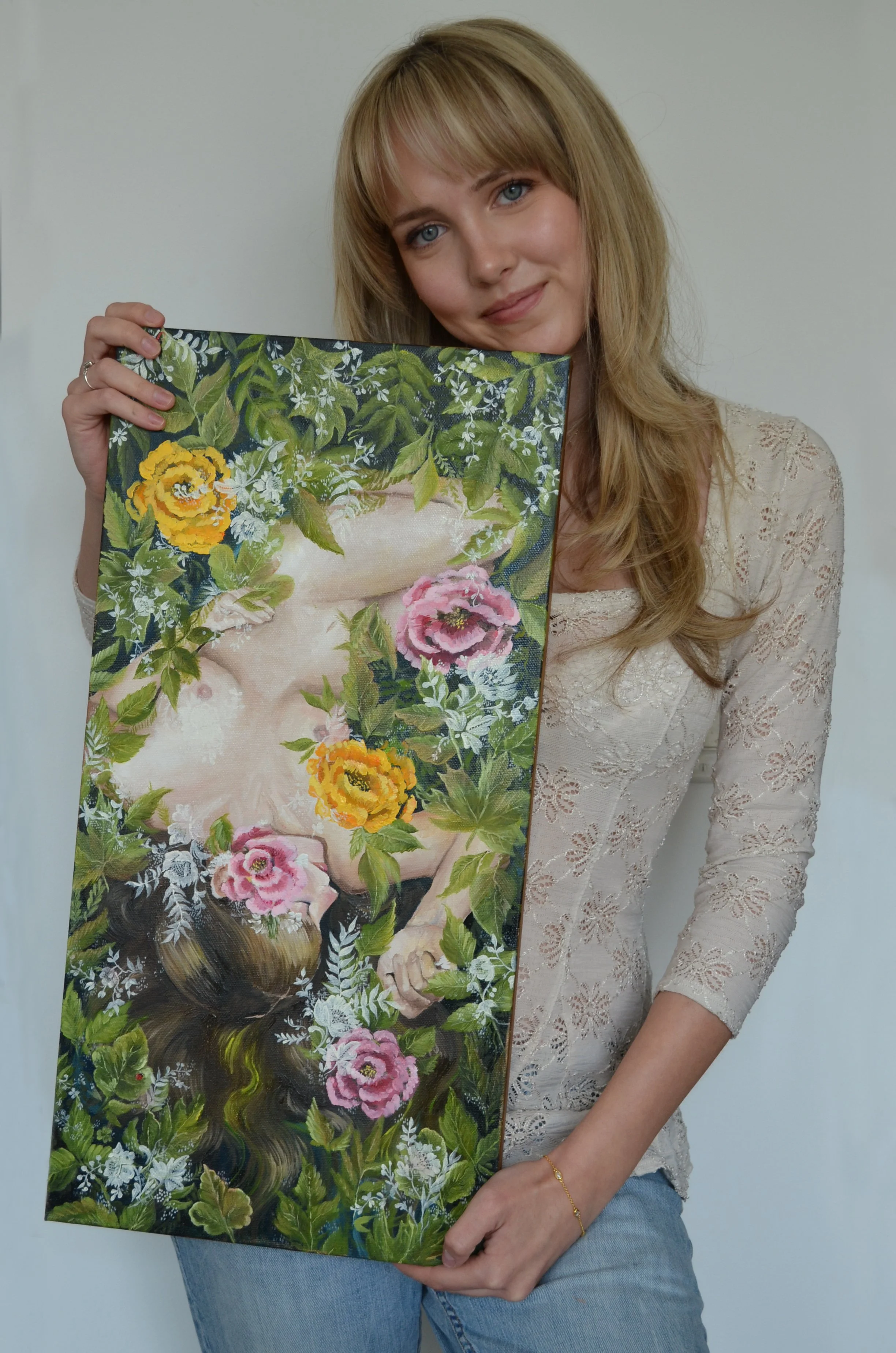 A woman with blonde hair, wearing a lace long sleeve top and jeans, holding a colorful floral painting of a woman surrounded by leaves and flowers.