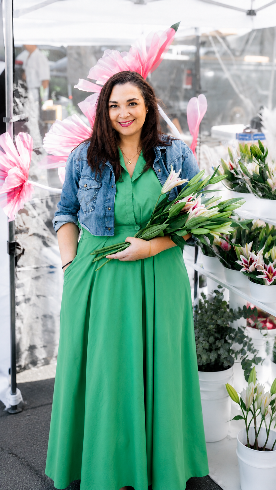 Woman with dark brown hair, wearing a green dress and denim jacket, holding white lilies at an outdoor flower market.