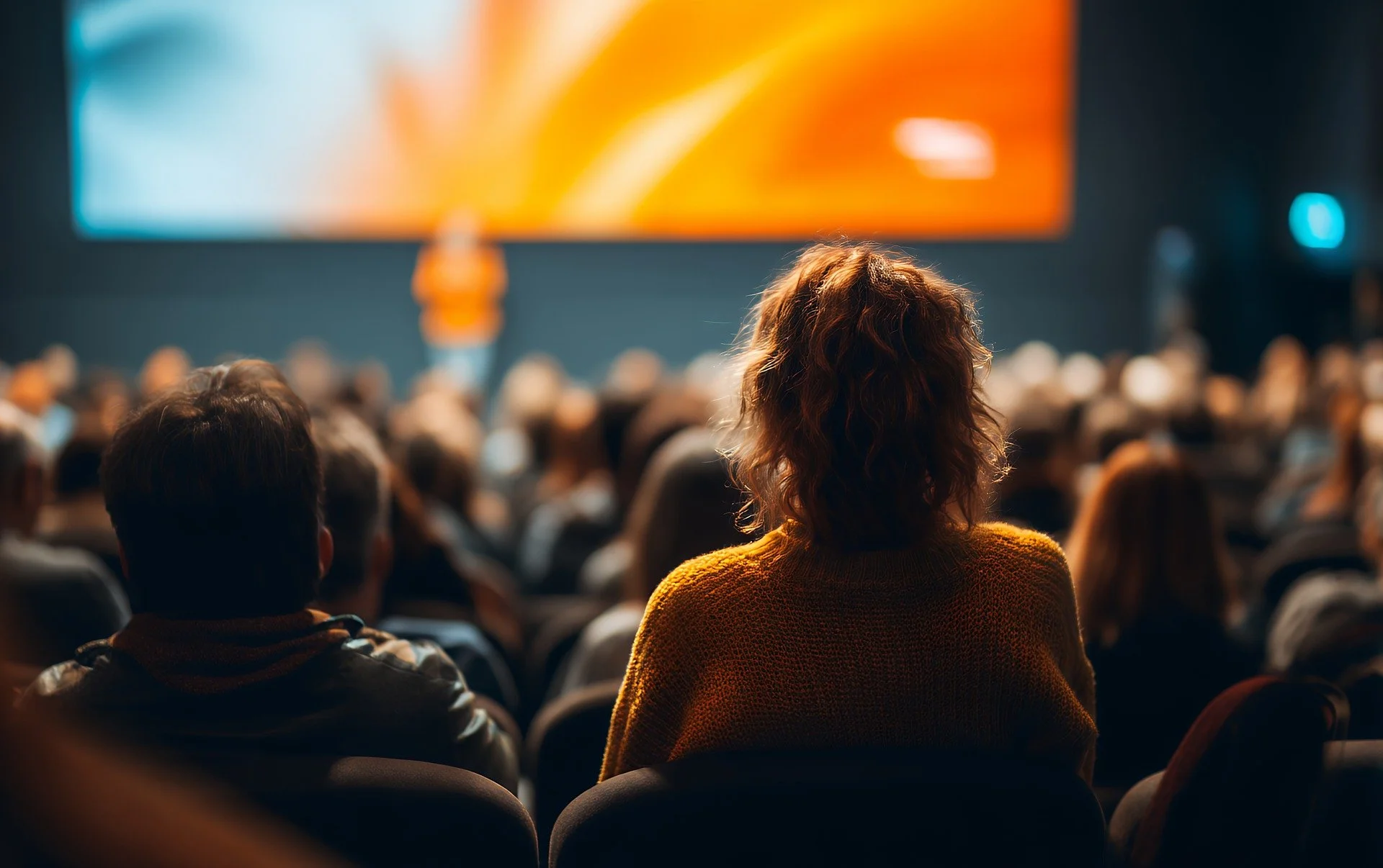 Audience attending a conference or presentation in a dimly lit room, with a large screen displaying an abstract orange and white background.
