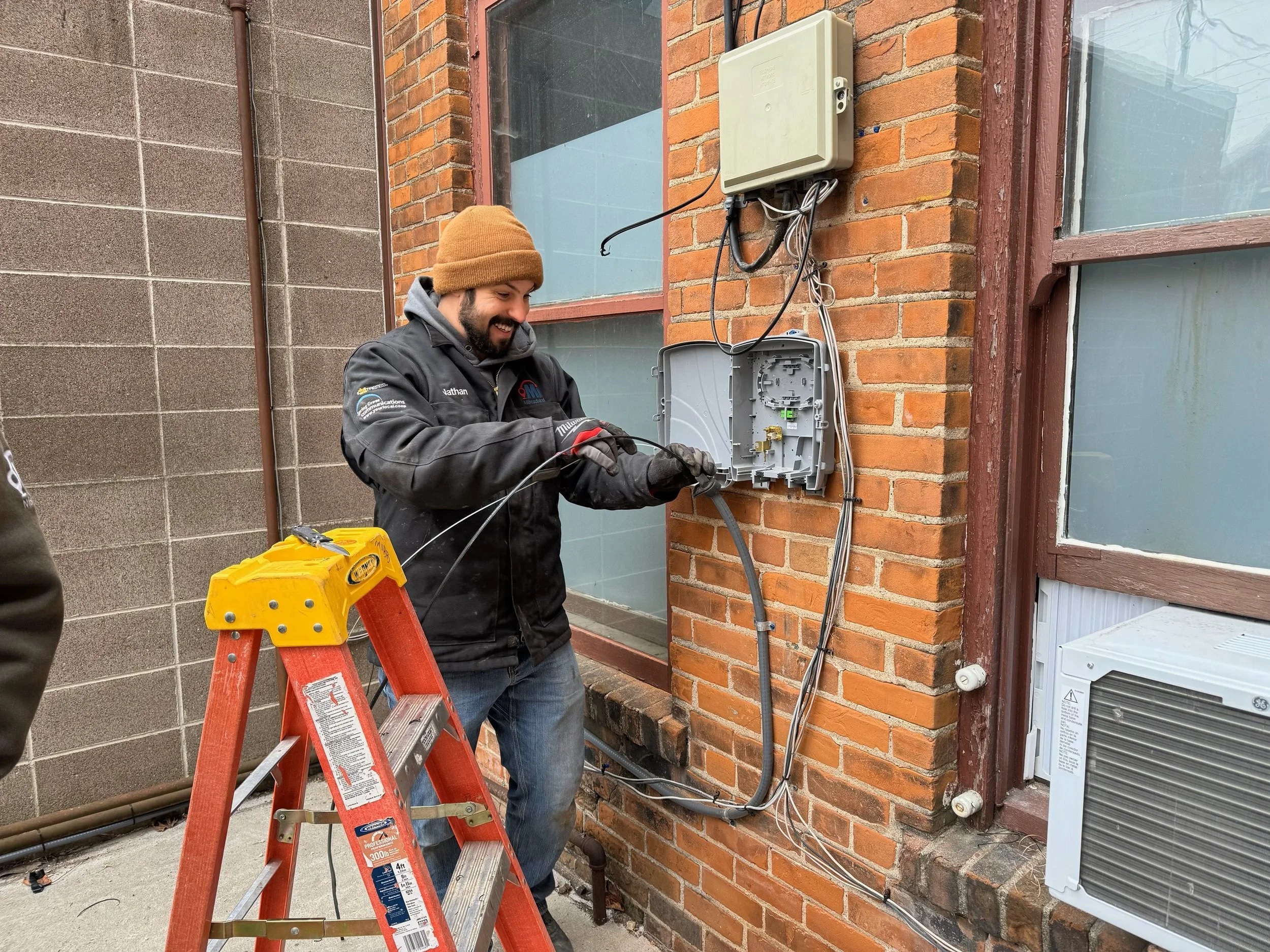 A man with a brown beanie smiling and working on outdoor electrical wiring next to a brick building, using a red ladder and yellow equipment.