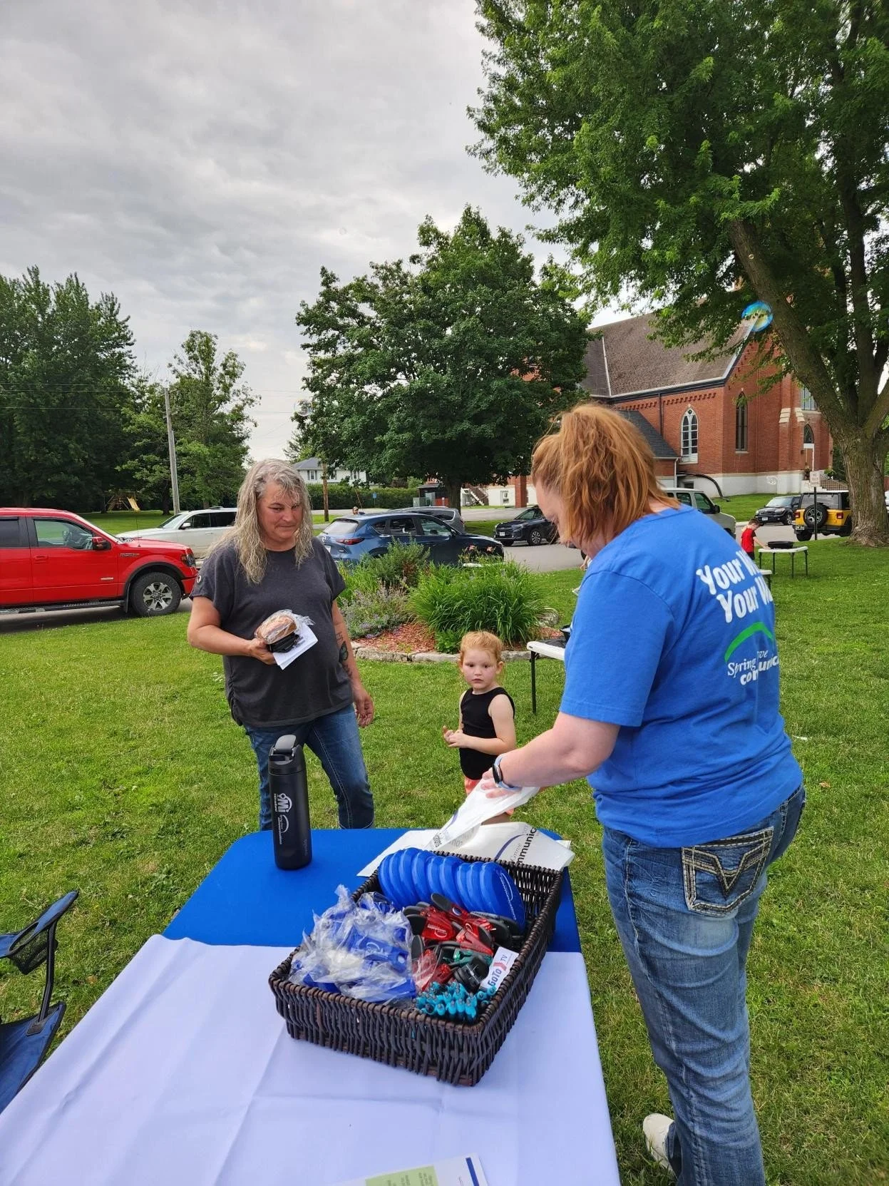 A woman with red hair wearing a blue shirt is organizing items on a table outdoors. Another woman with long blonde hair, wearing a dark shirt and jeans, holds a plastic bag with food. A young girl with light hair and a black shirt looks on. The setting is a grassy area with trees, parked cars, and a brick church in the background.